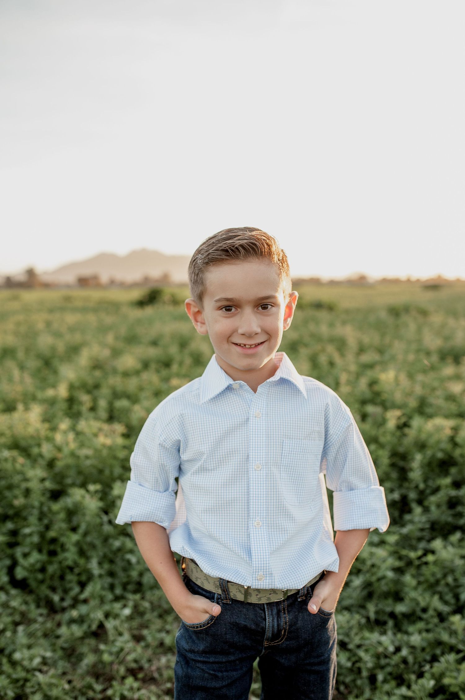 A young person stands in a green field at sunset wearing a light blue button-down shirt and jeans.