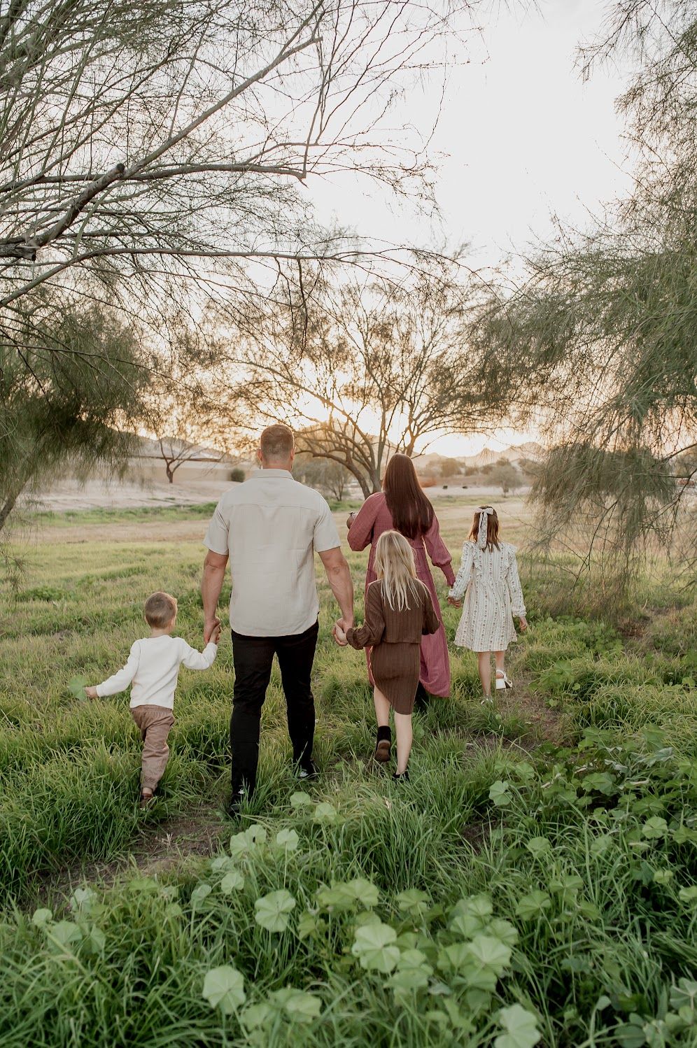 Family of five walking hand-in-hand through a lush green meadow at golden hour sunset.