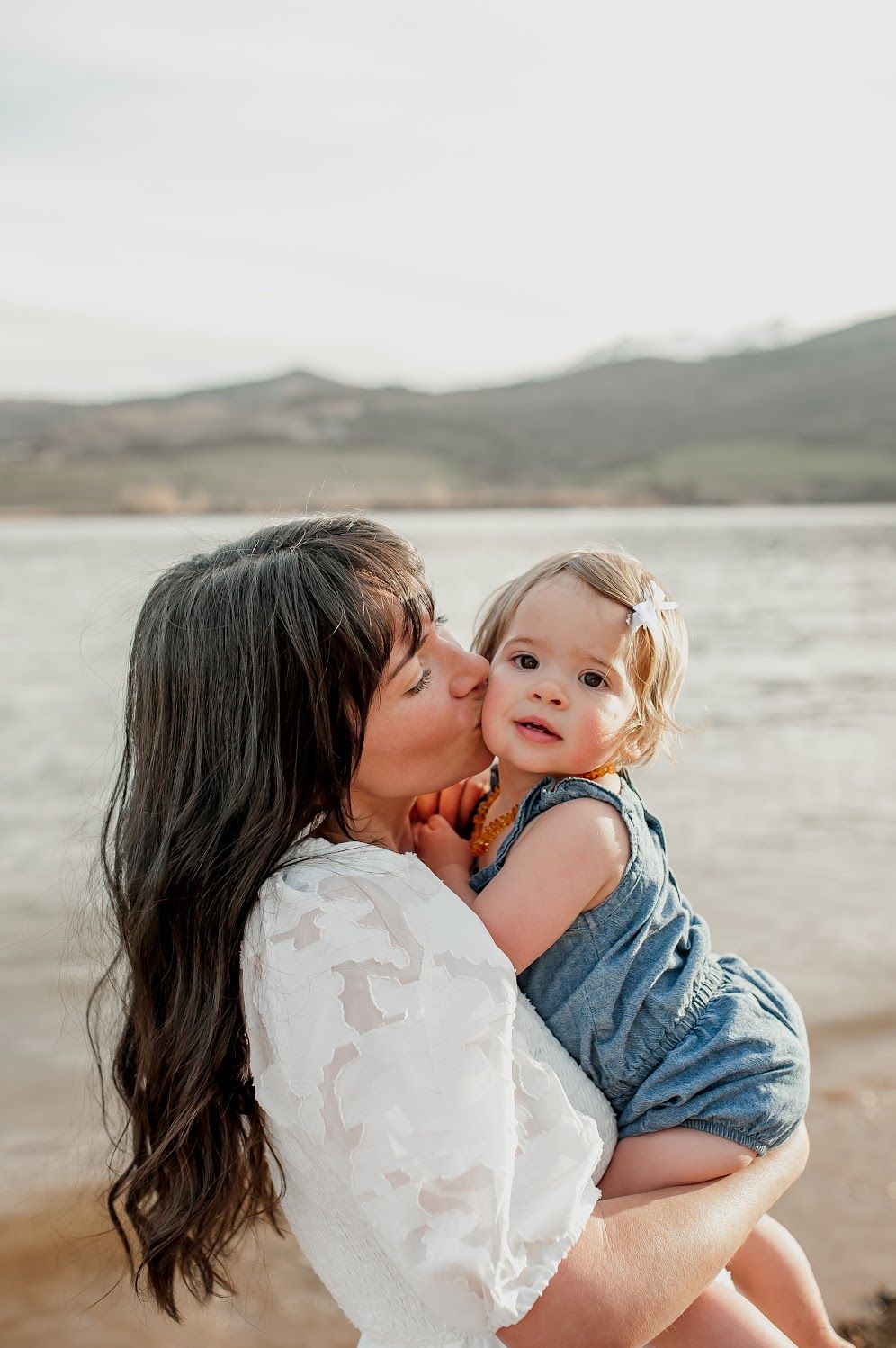 A mother with dark hair kisses her toddler girl in a denim romper near a lake with mountains behind.