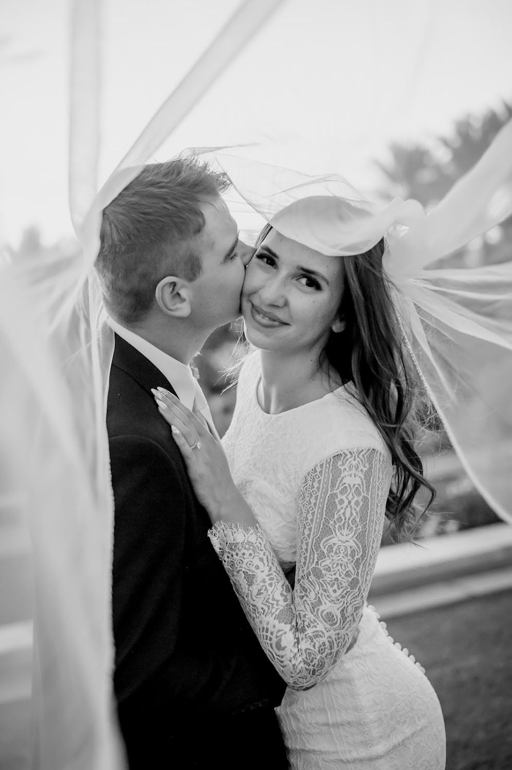 Black and white wedding photo of groom kissing bride on cheek under flowing veil.