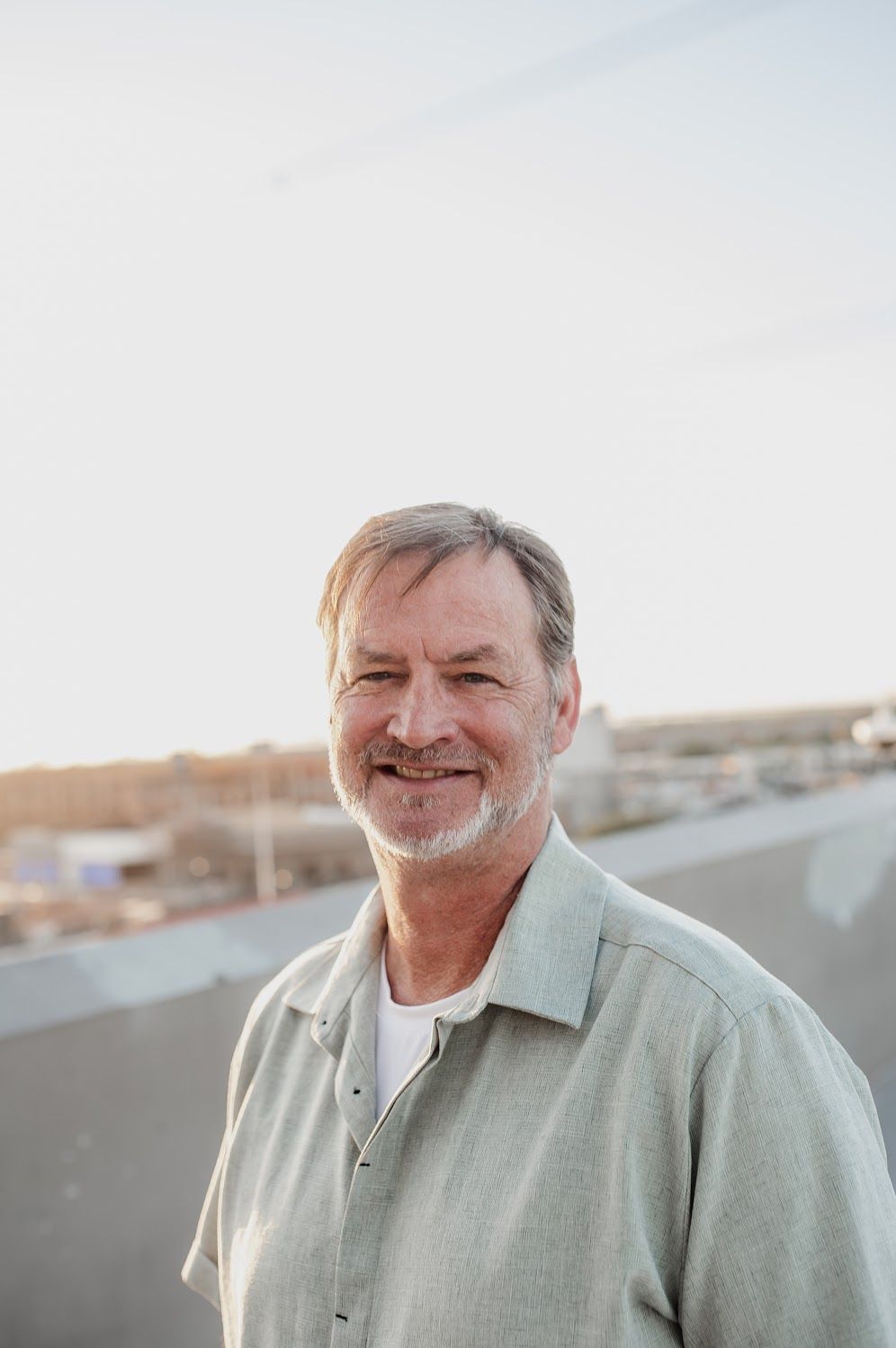 Smiling middle-aged man with gray hair and beard wearing a light green shirt outdoors at sunset.