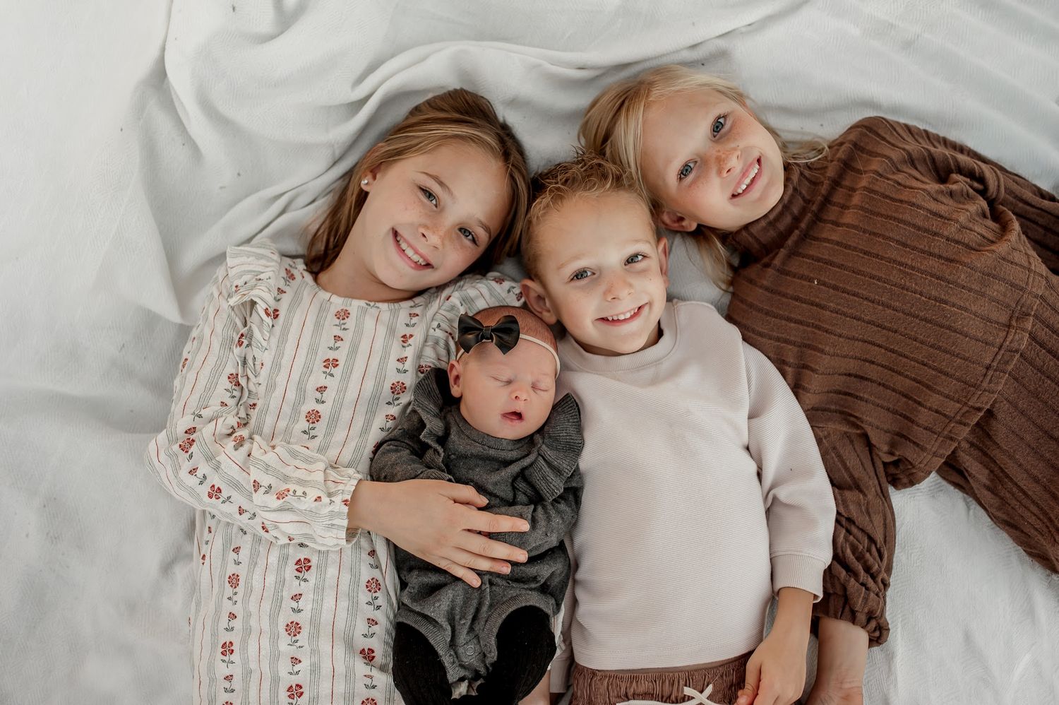 Three smiling siblings lying on white bedding, two older girls in dresses flanking a toddler boy holding a newborn baby.