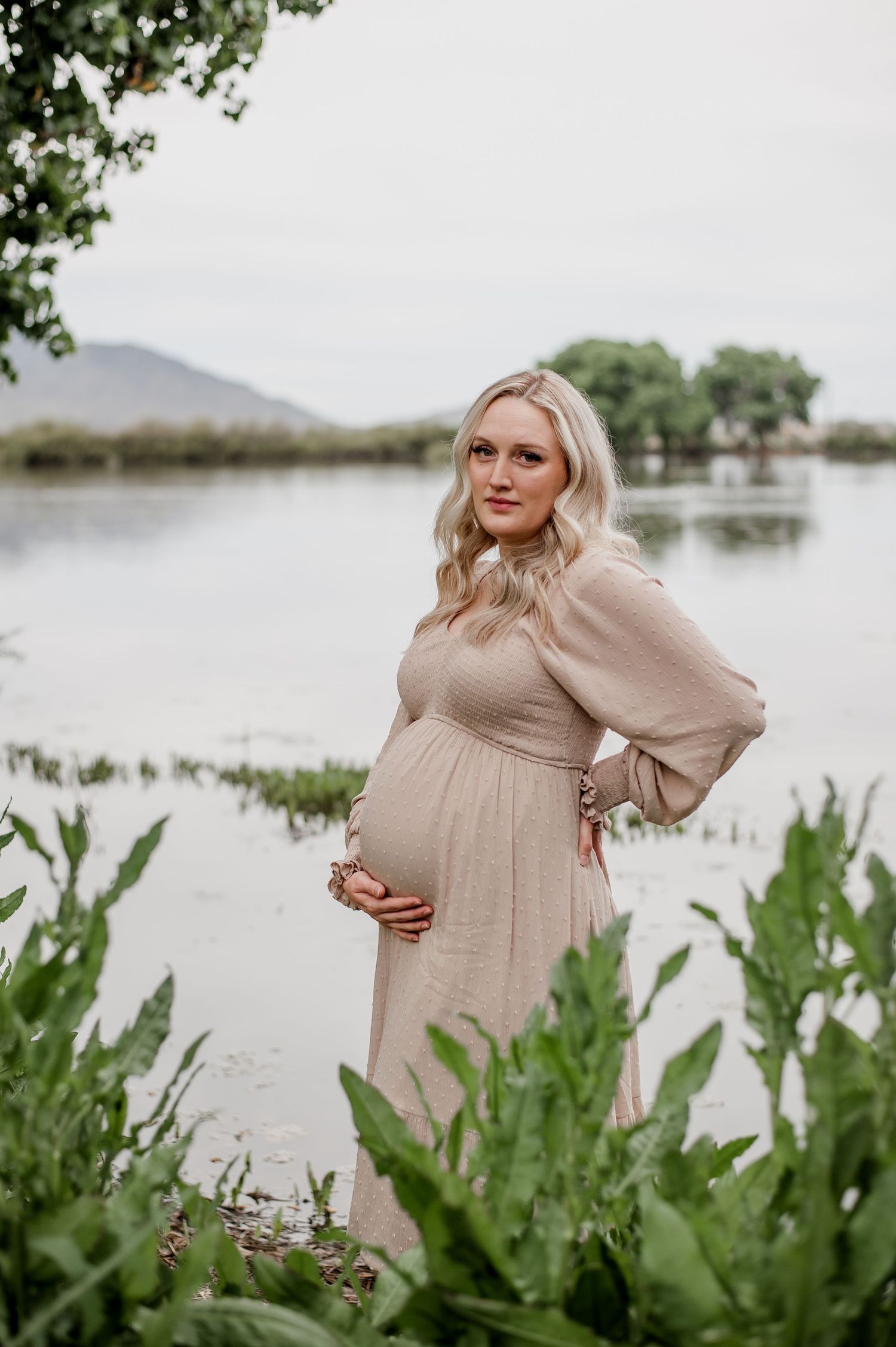 Expectant mother in beige dress poses elegantly by lakeside surrounded by lush vegetation.