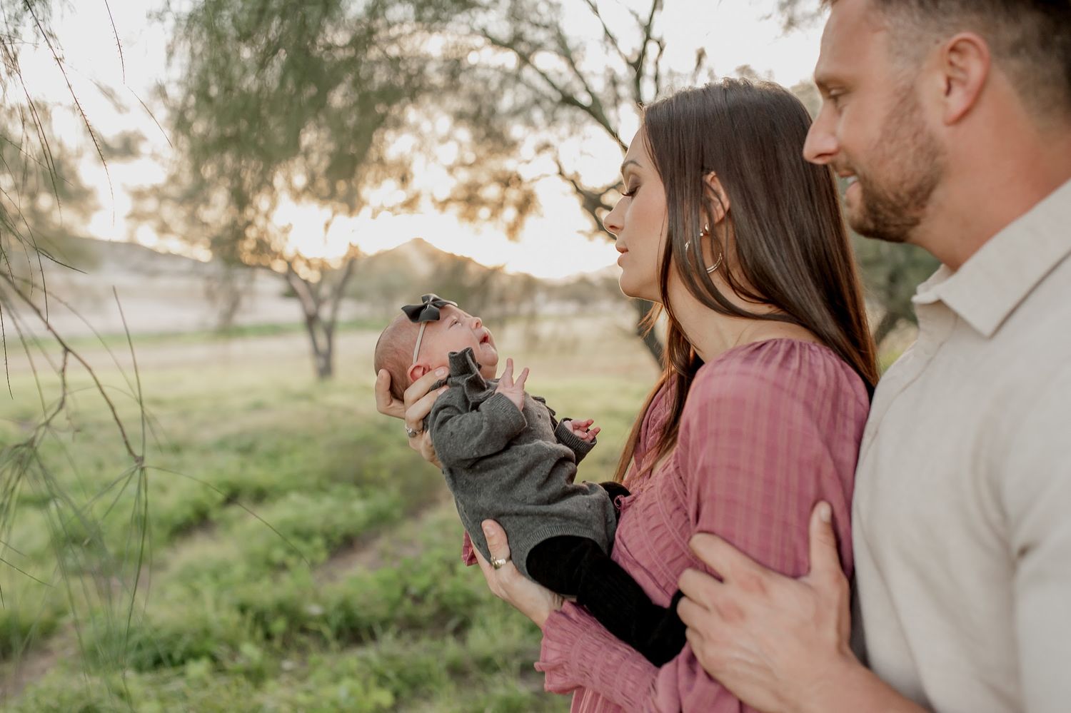 Parents embrace outdoors in a sunlit green field while the mother gently holds and kisses their newborn baby in a family portrait.
