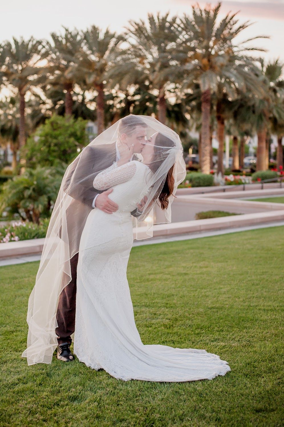 Bride and groom sharing a romantic kiss under a flowing veil on green lawn with palm trees at sunset.
