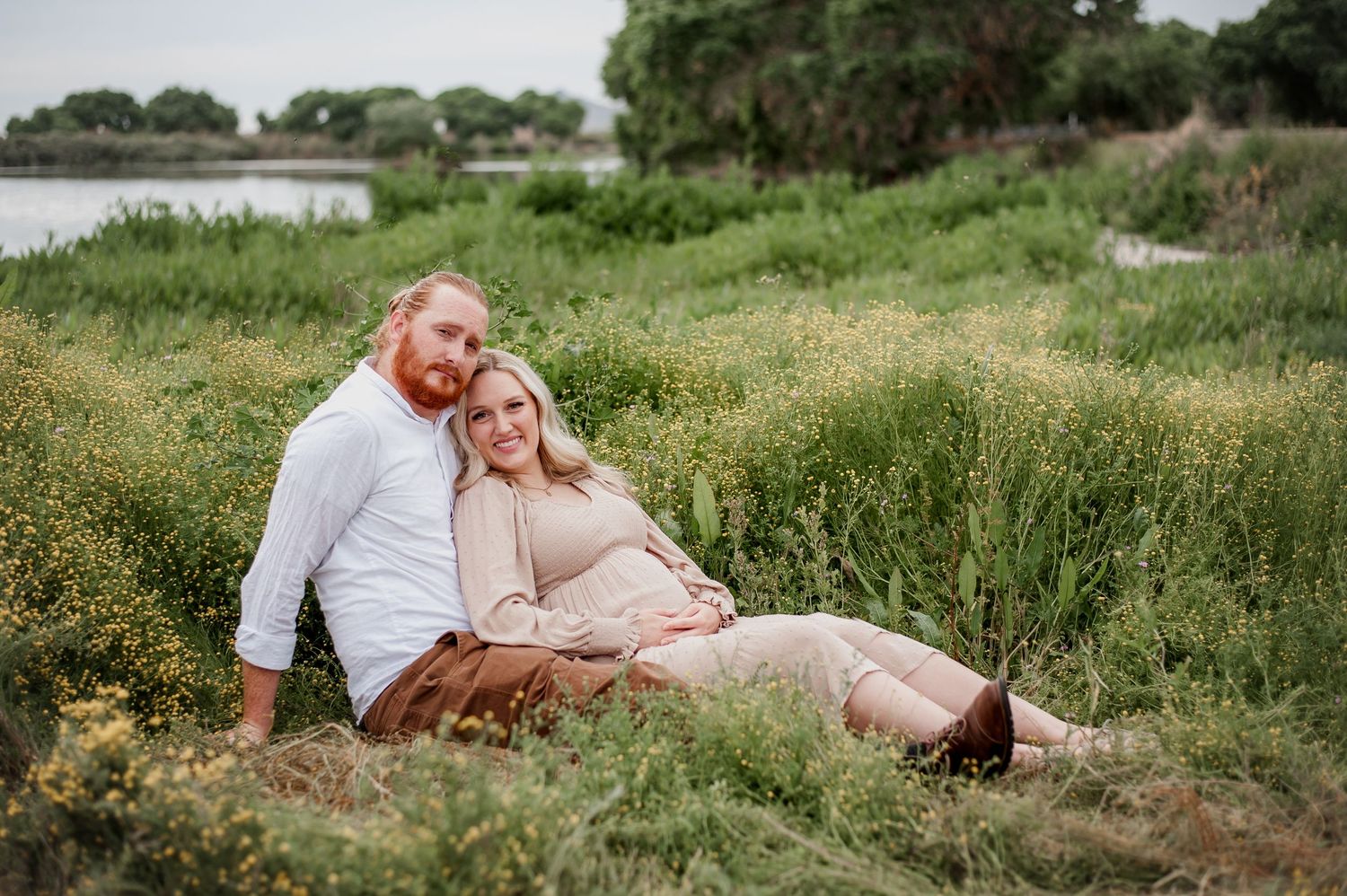 A couple sits together in a grassy meadow near a lake on a sunny day.
