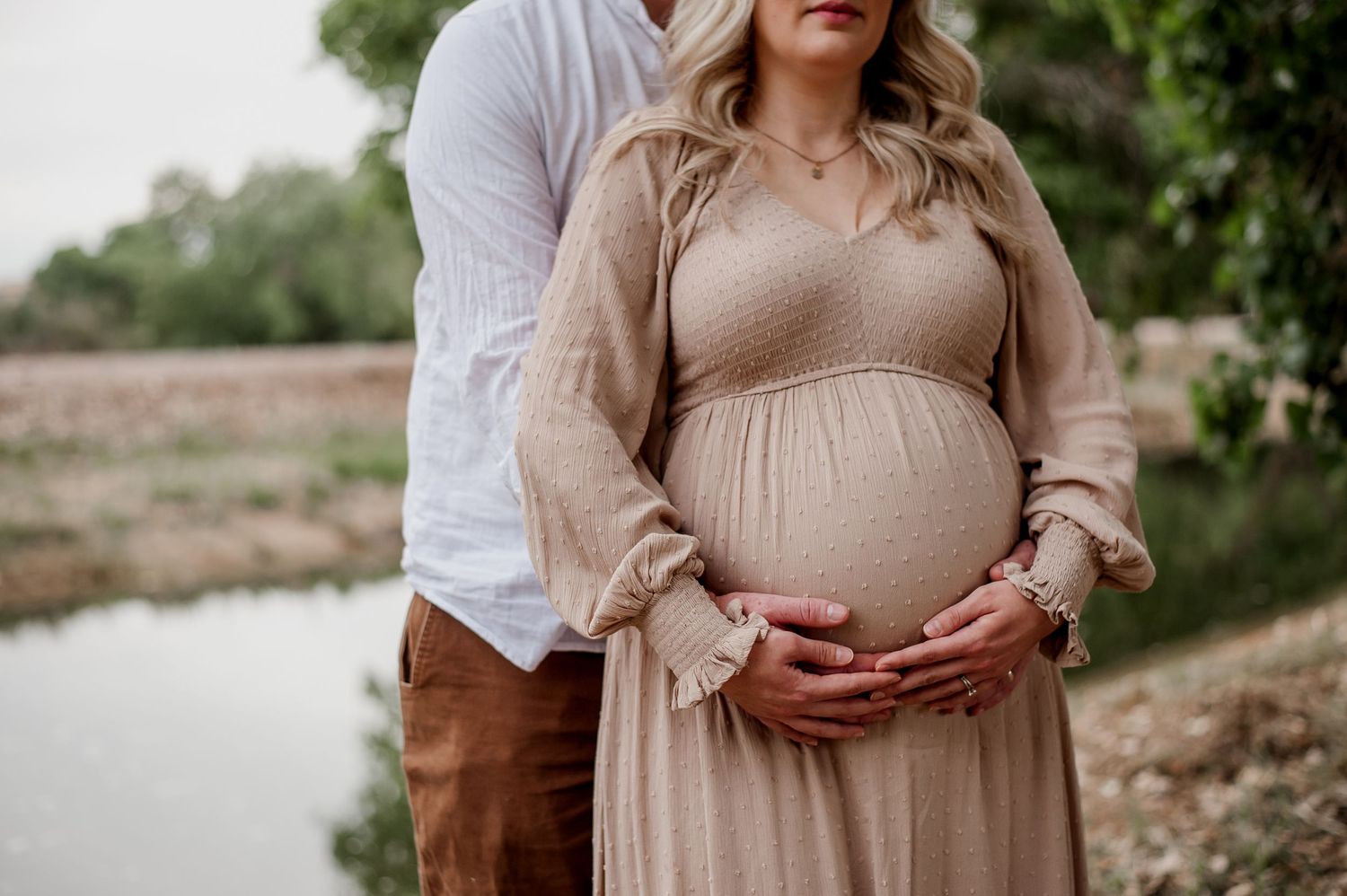Intimate maternity photoshoot by a lake with couple in neutral tones embracing amidst natural greenery.