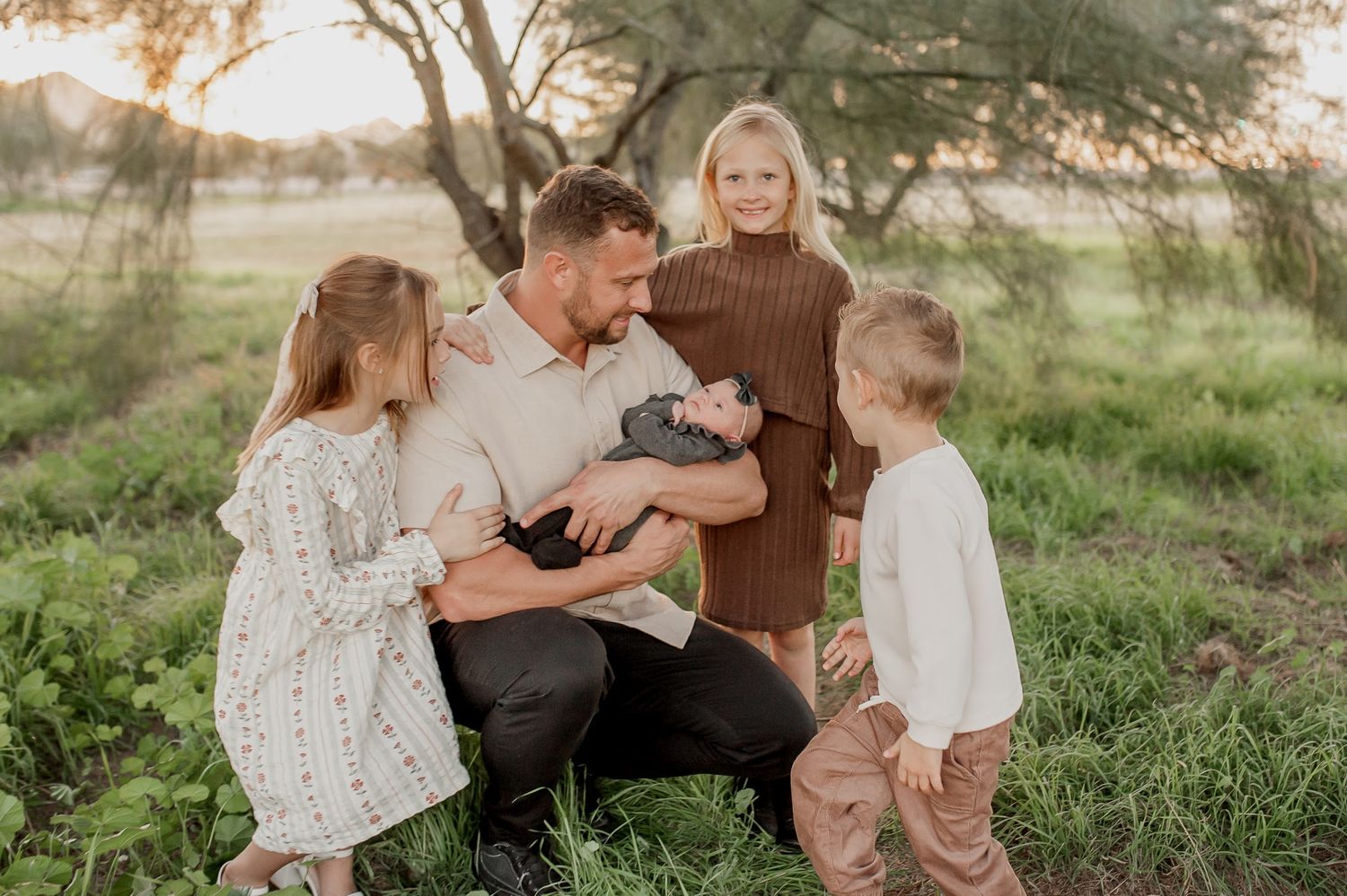 Family of five gathered outdoors at sunset, father kneeling and holding newborn baby surrounded by three young children.