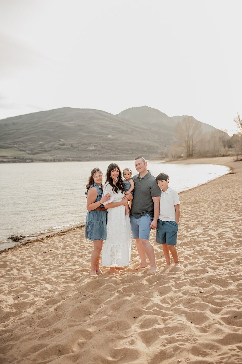 Family of five posing together on a sandy beach with mountains and lake in the background on a sunny day.