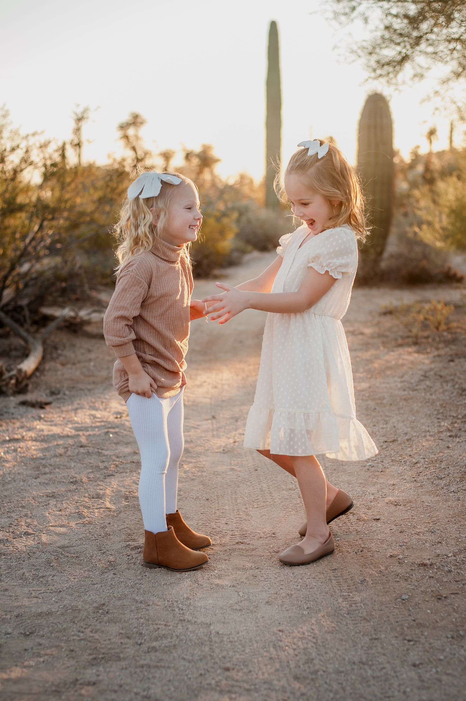 Two young girls in matching hair bows share a sweet moment on a desert path surrounded by saguaro cacti.