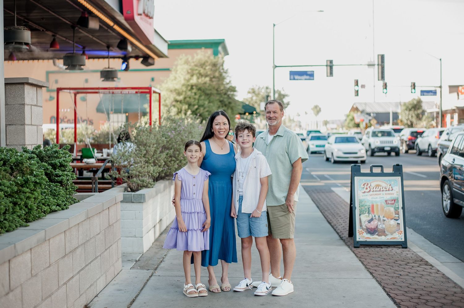 Family of four posing on a sunny sidewalk in a suburban shopping district with palm trees and traffic lights.