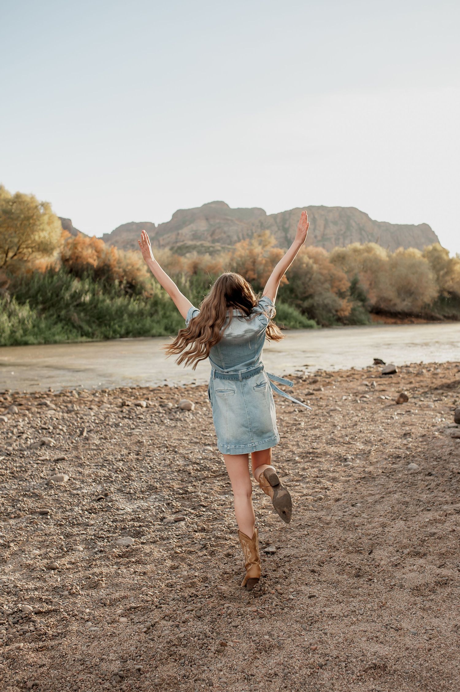 Person in denim shorts and boots dances joyfully on a rocky beach with majestic mountains in the background.