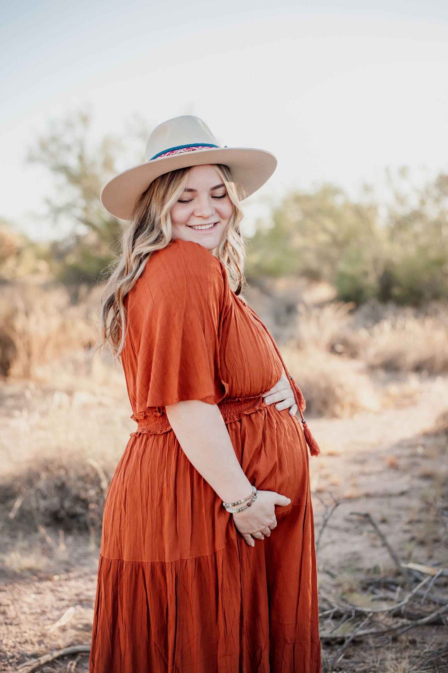 A pregnant woman in a rust-colored maxi dress and wide brim hat poses in a desert landscape during golden hour.