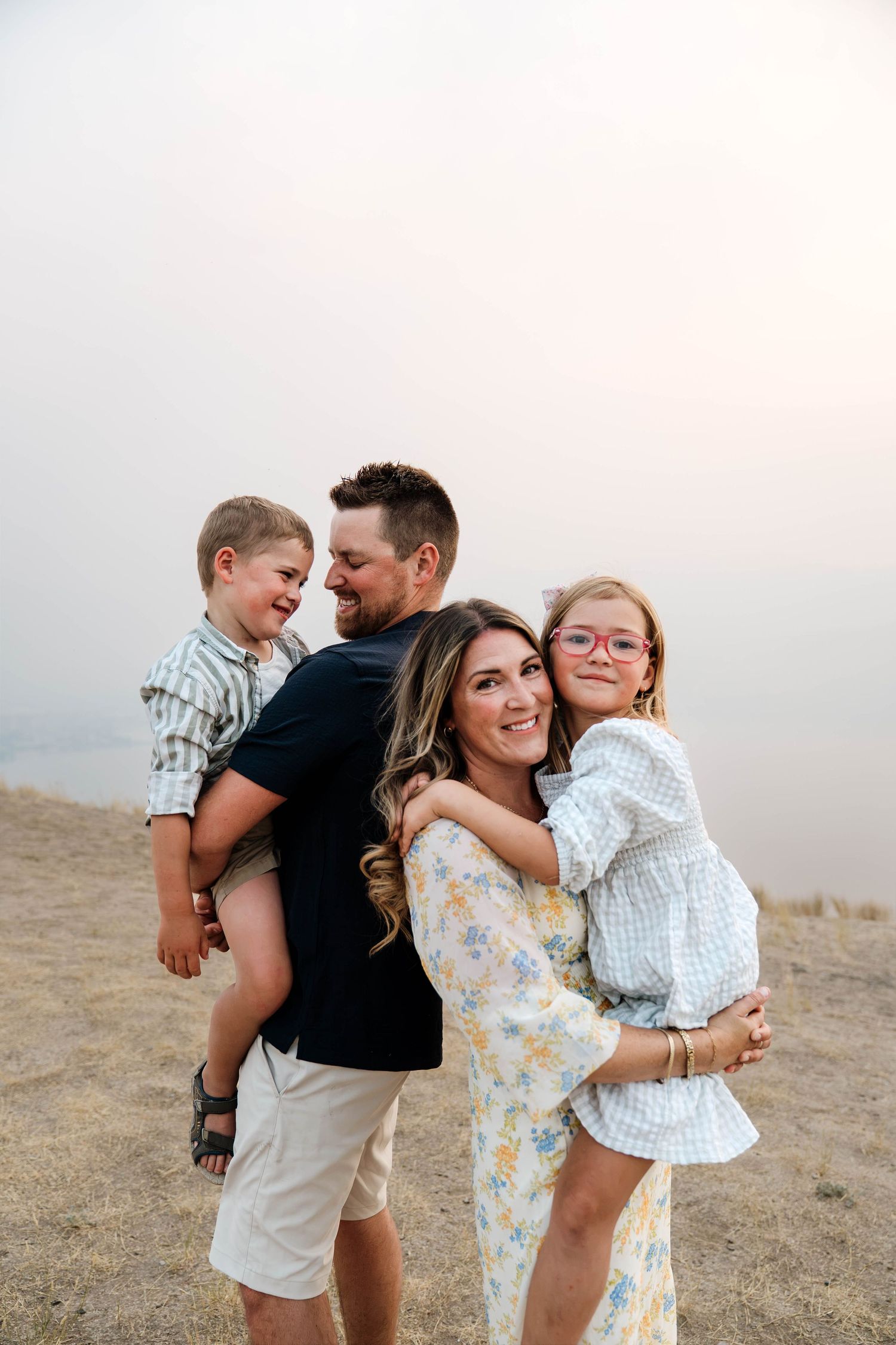 A happy family of four poses outdoors on sandy terrain, with parents holding their two young children while smiling.