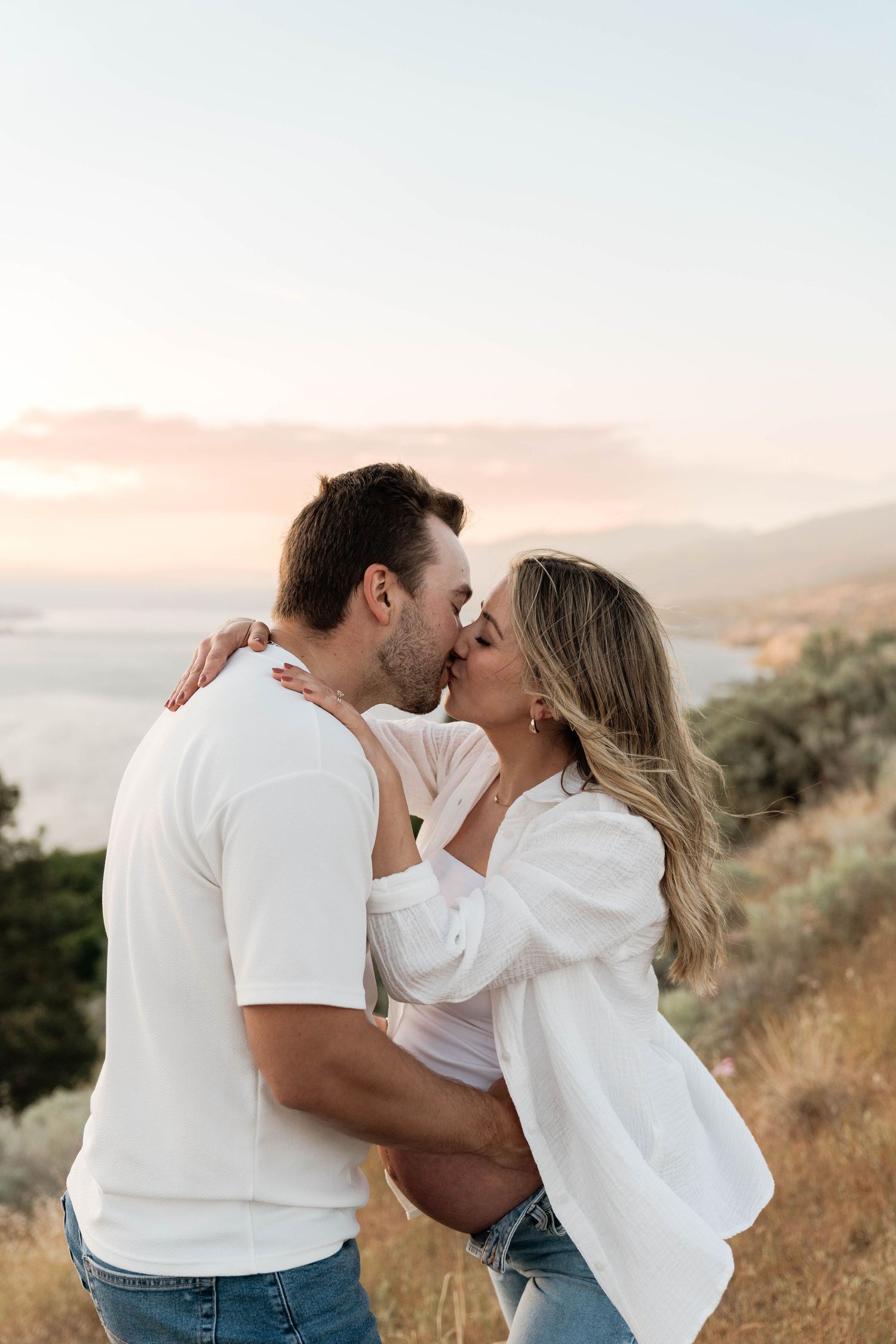Couple kissing outdoors on a hillside with a scenic mountain lake visible in the soft evening light in Penticton