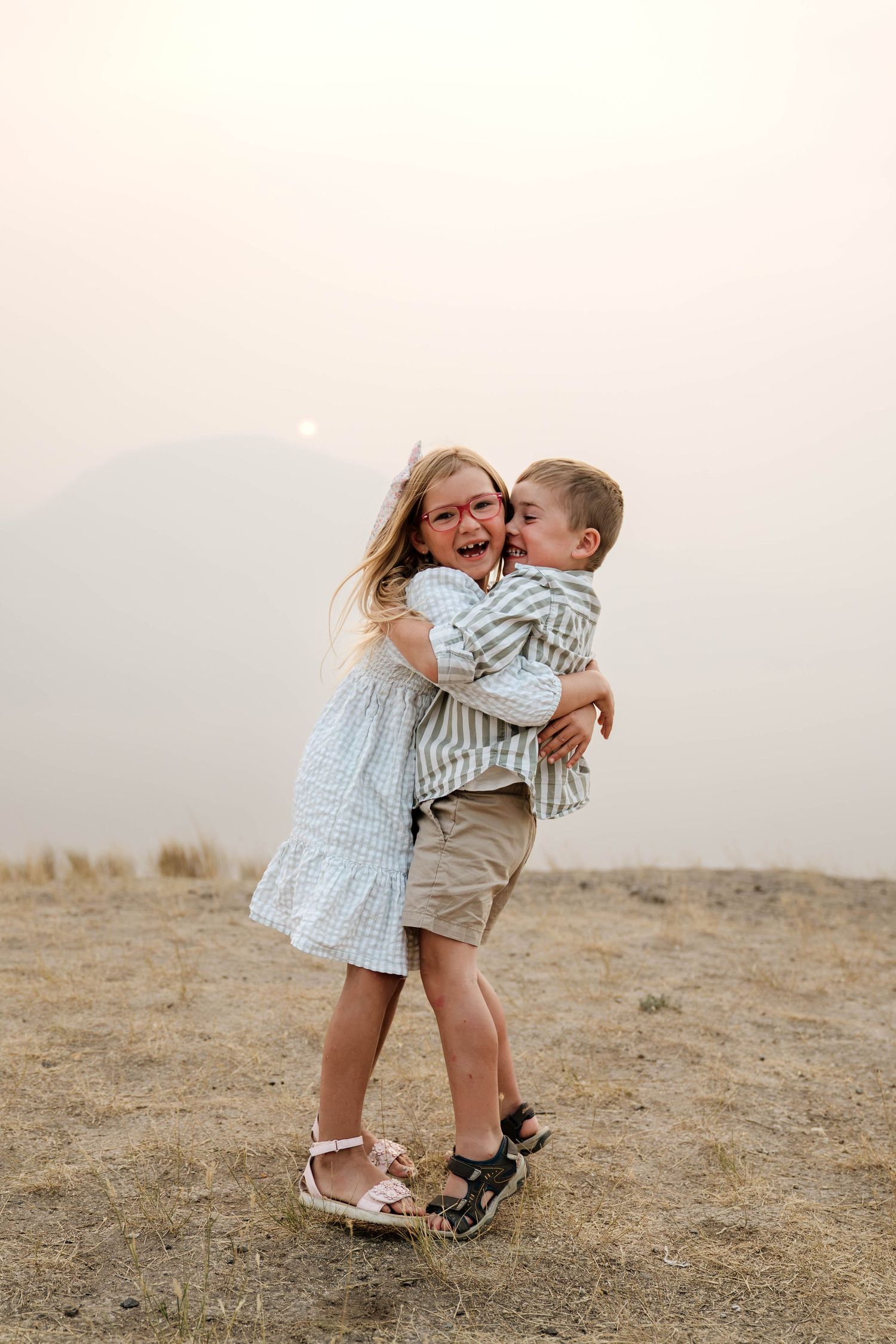 Two young children hugging and laughing on a hillside, girl in blue dress and boy in striped shirt, misty mountains behind.