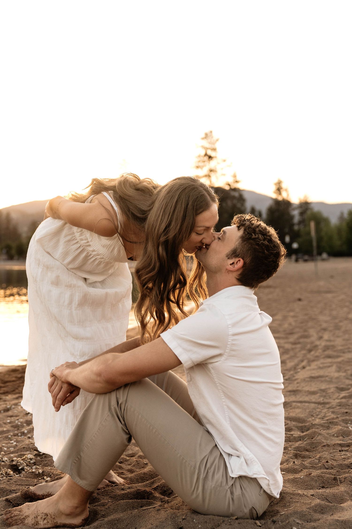 Couple sharing a romantic kiss on a sandy beach at golden hour sunset, woman in white dress leaning toward seated man.
