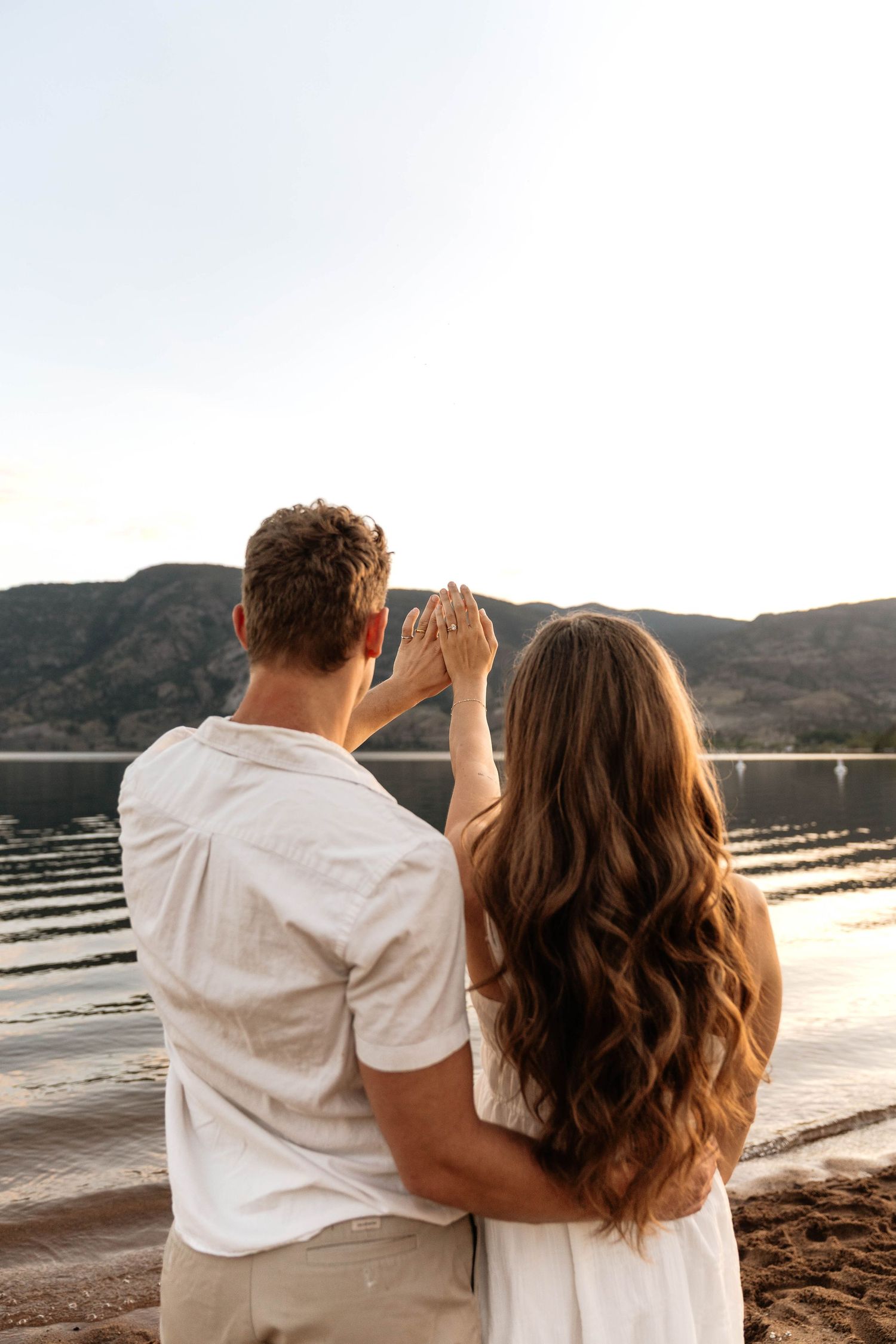 Couple holding hands facing a scenic mountain lake at sunset, viewed from behind, wearing white outfits.