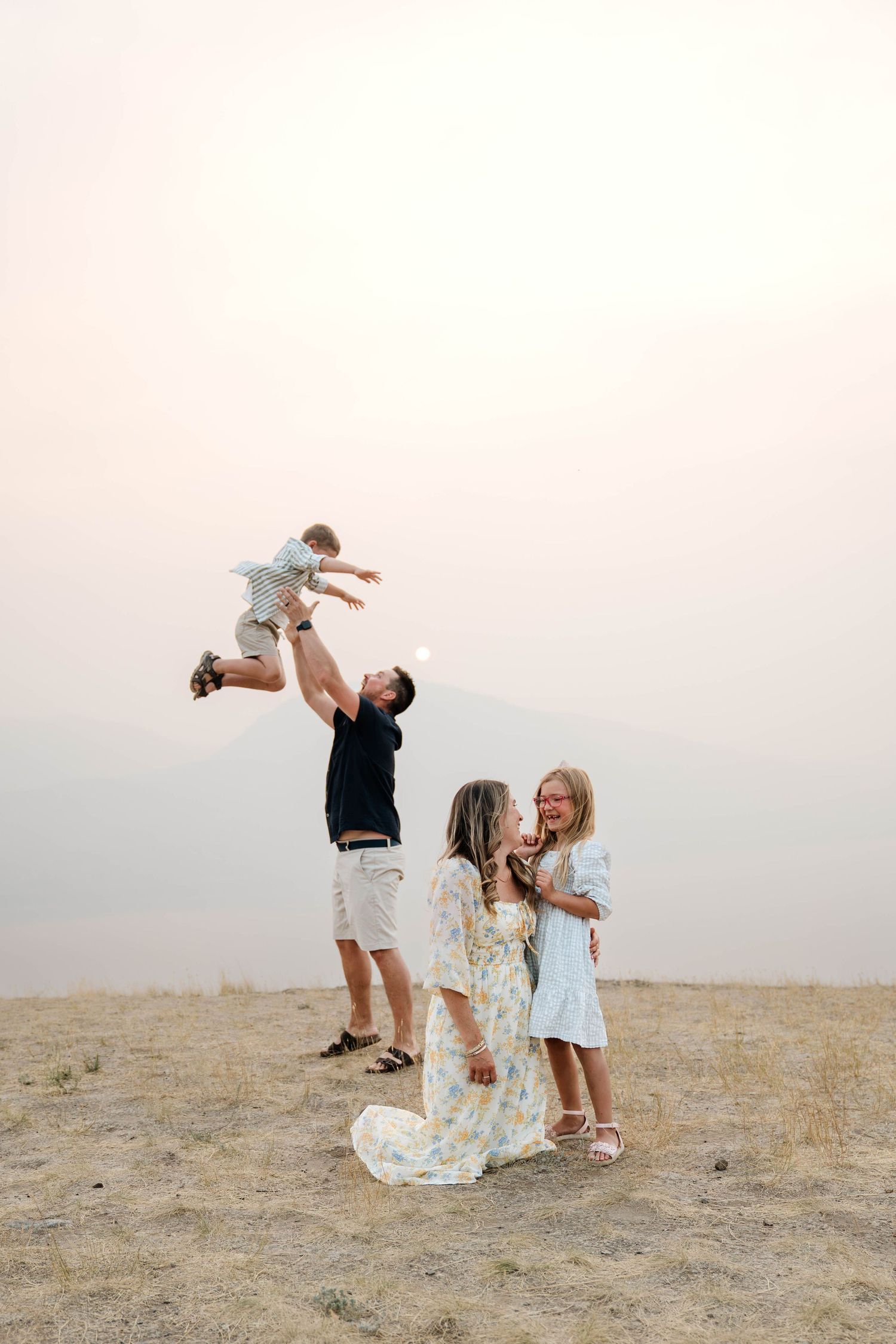 Family photo with dad tossing baby in air while mom and daughter embrace outdoors at sunset.