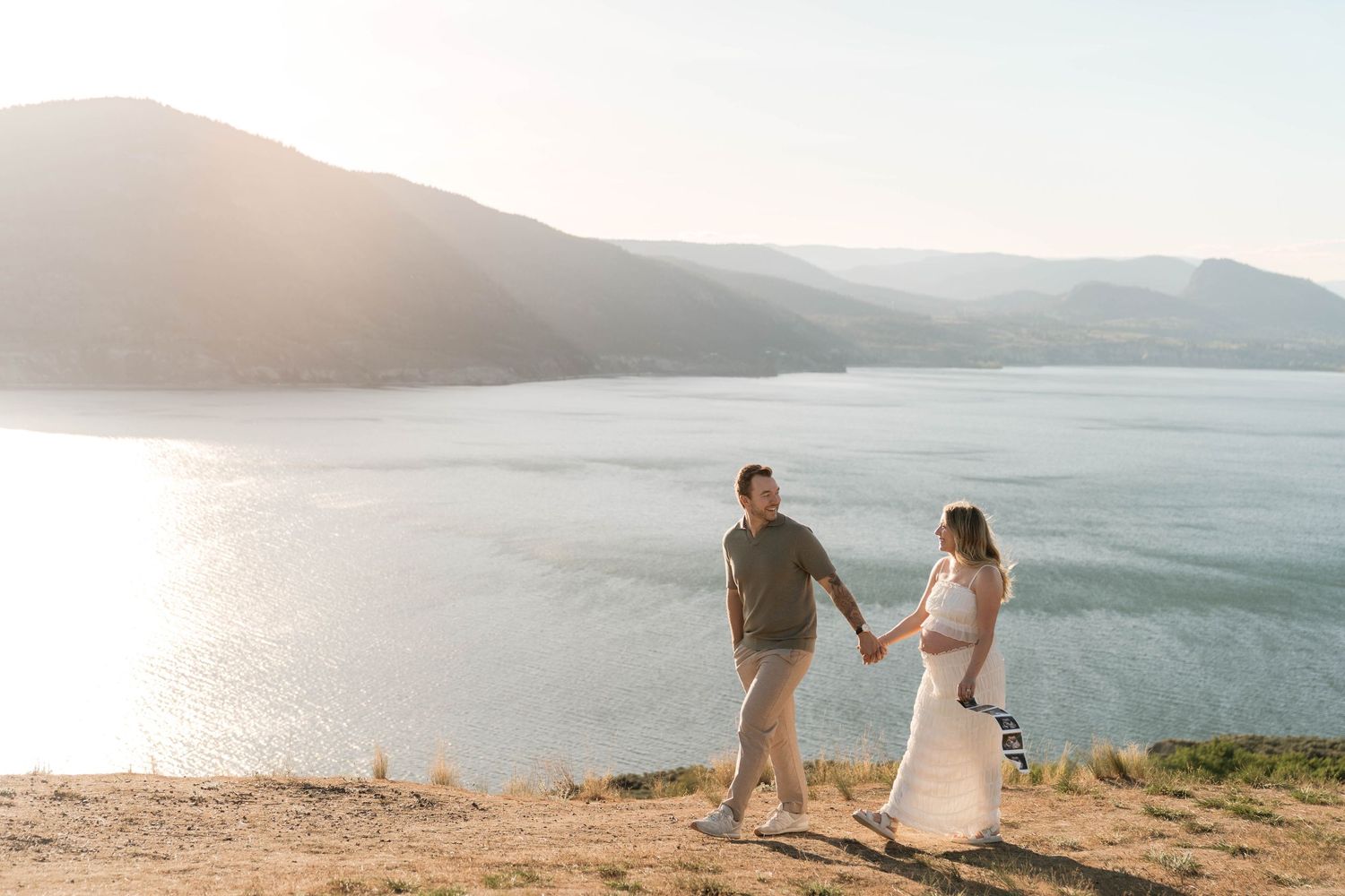 Couple holding hands walking on a sunlit hillside with a scenic lake and mountains in the background.