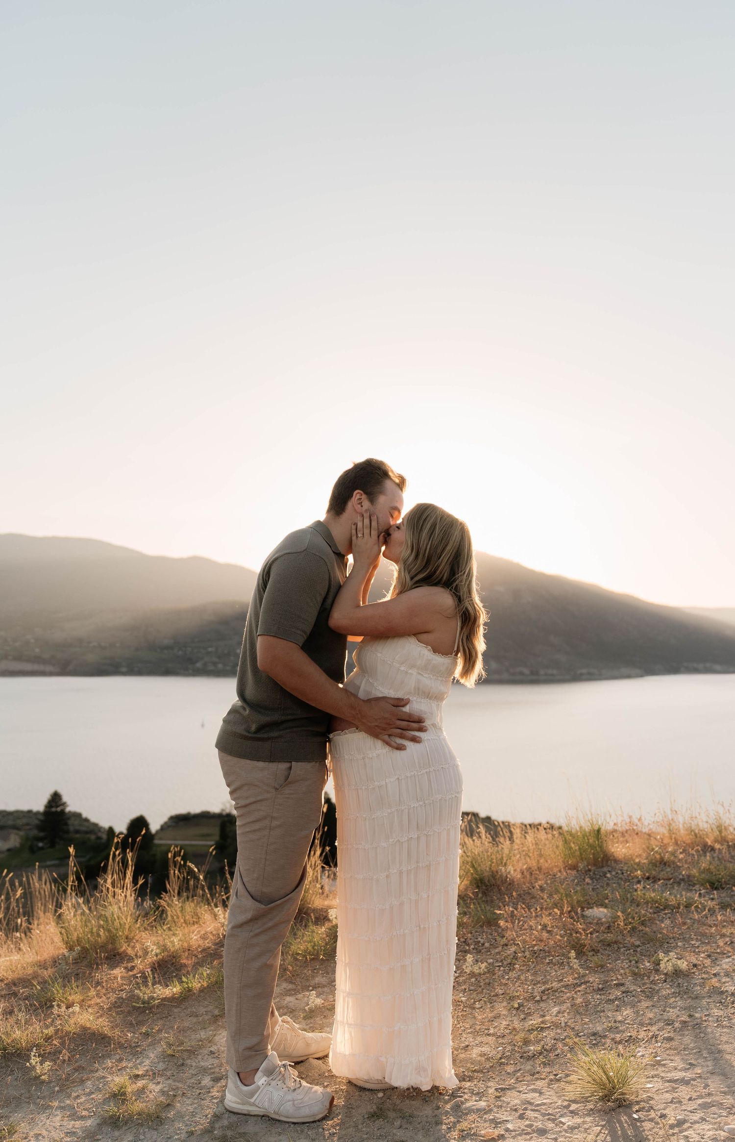 Couple sharing a romantic kiss on a hilltop overlooking a mountain lake during a golden sunset in Penticton