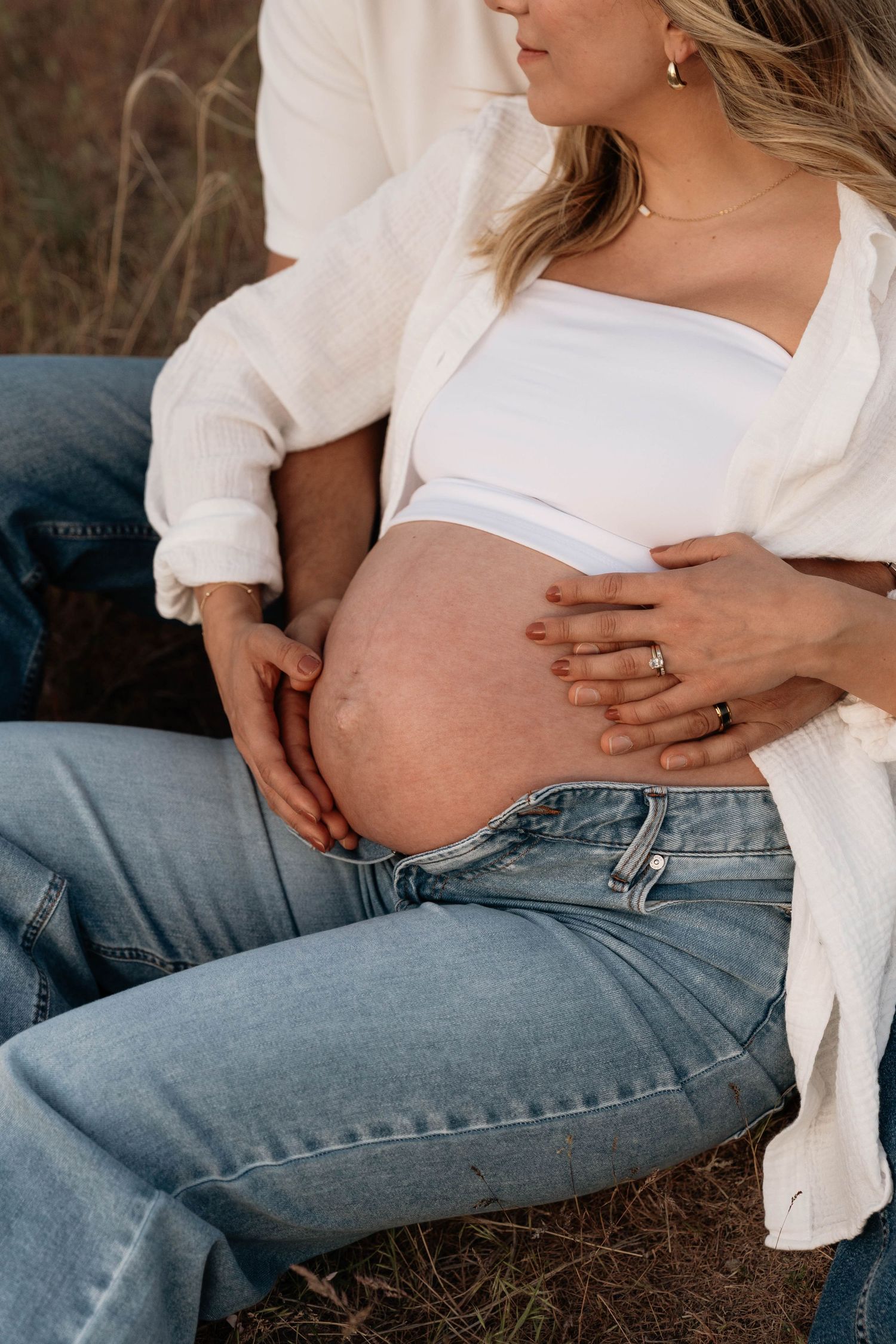 Pregnant woman in white top and jeans sits outdoors with partner, both cradling her bare baby bump.