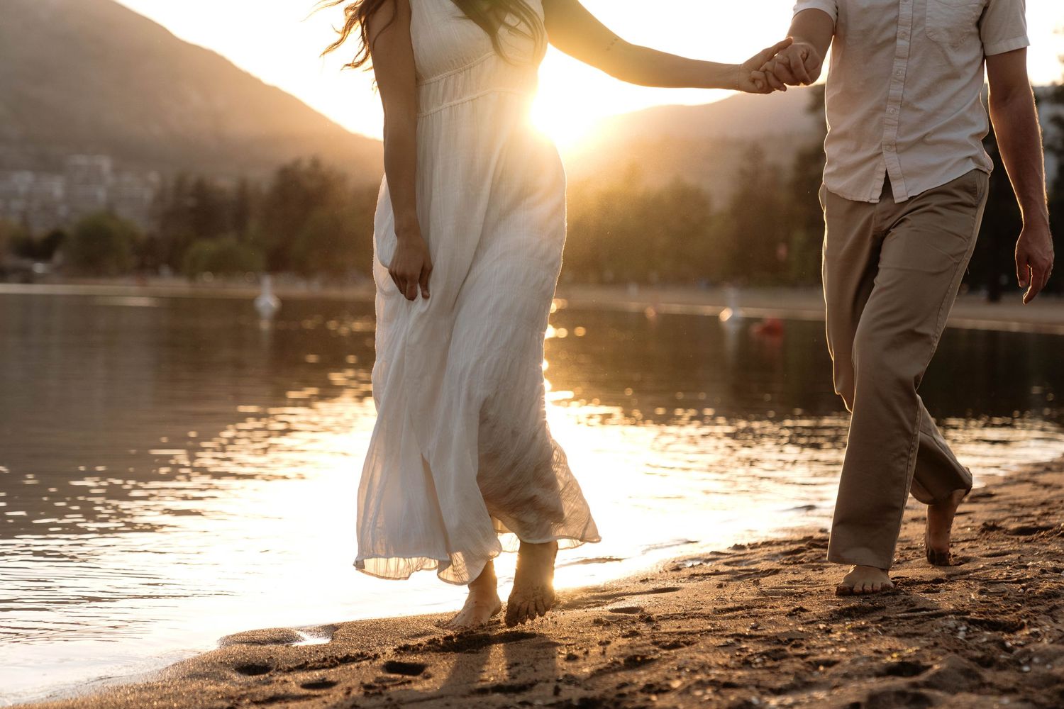 Couple walking barefoot on sandy beach at golden sunset, woman in white dress holding hands with man in casual attire.