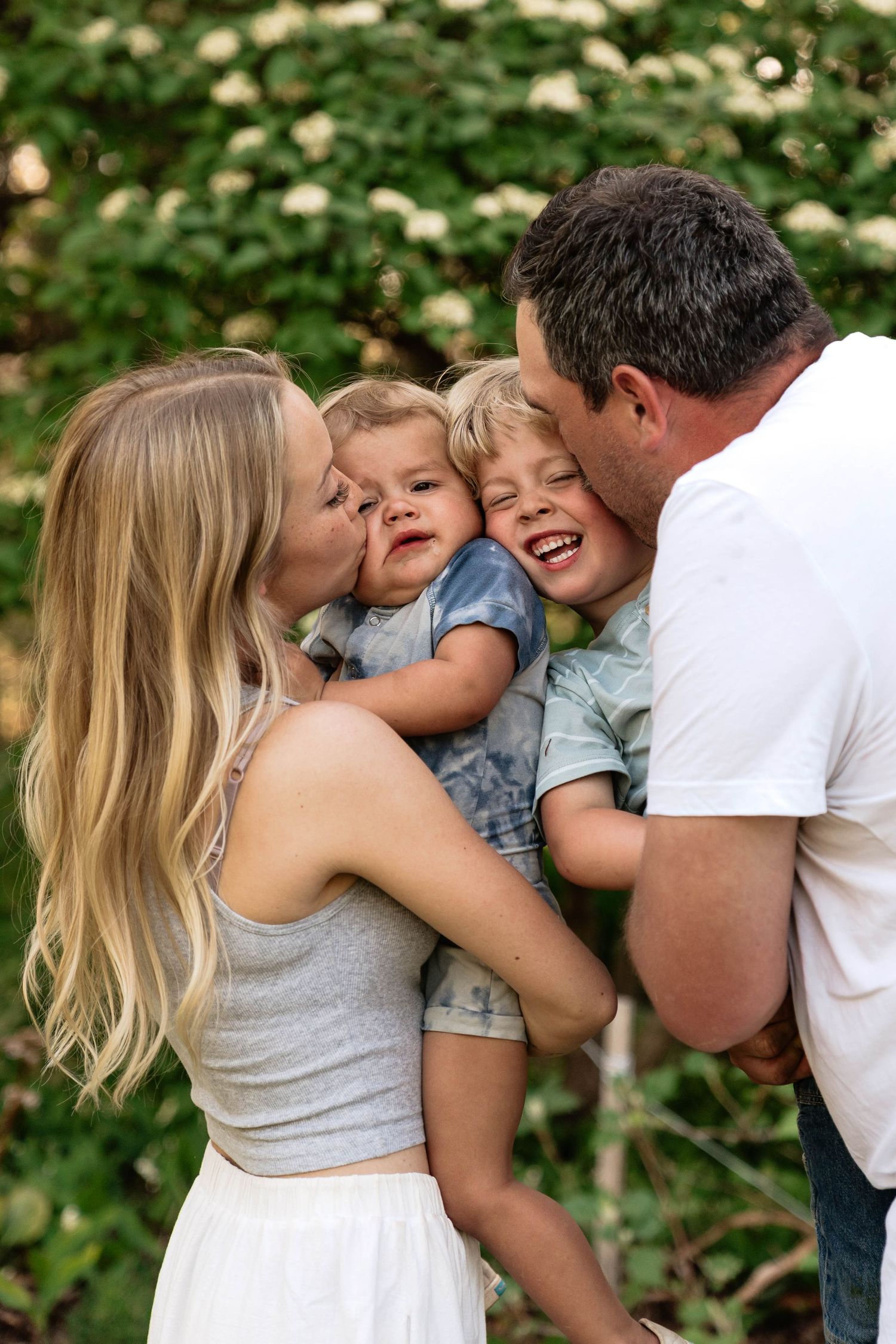 A happy family with two young boys shares kisses and laughter outdoors in front of blooming white flowers in Penticton