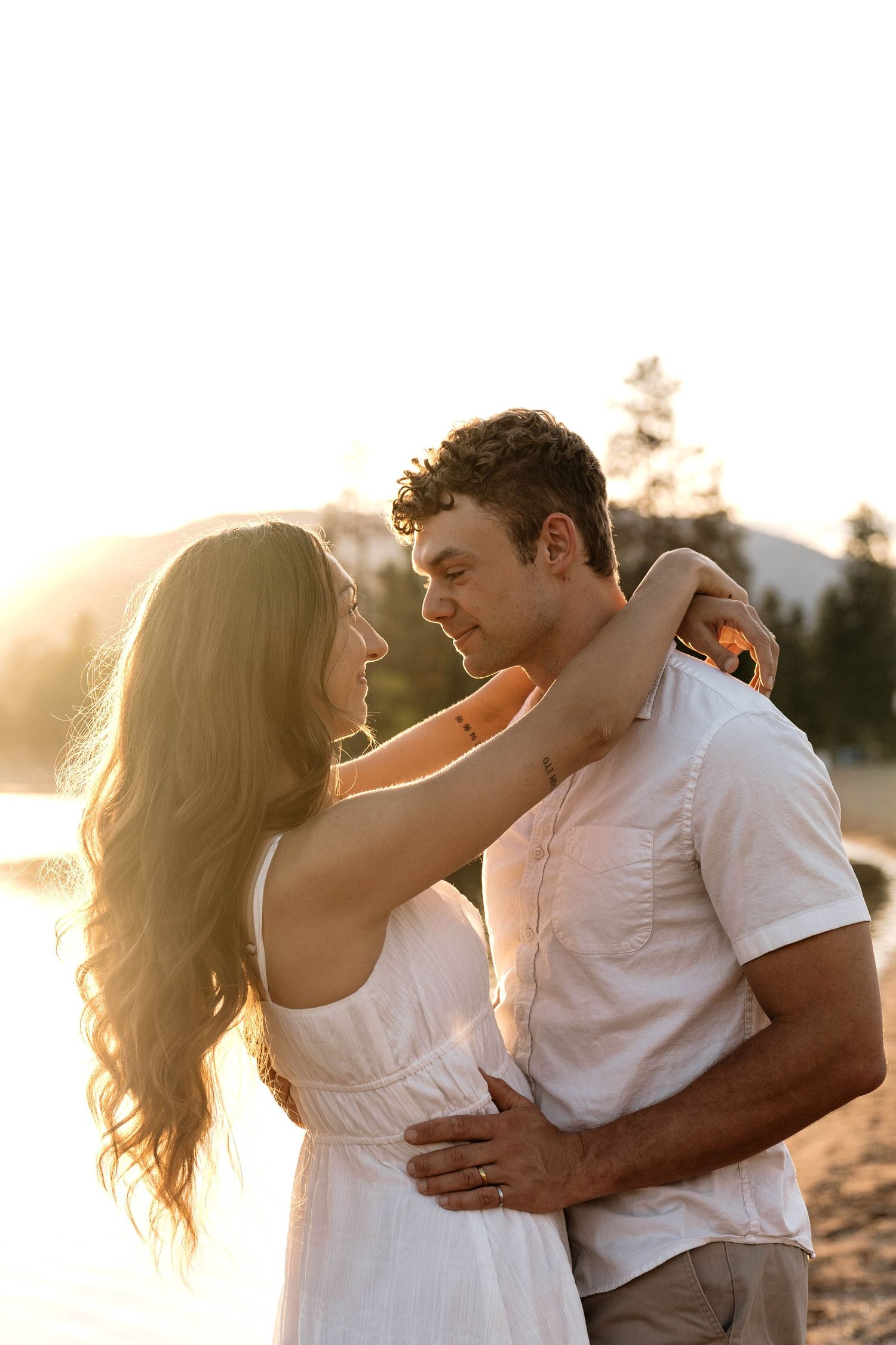 A couple embraces at golden hour sunset by a lake, the woman in a white dress with her arms around the man's neck.