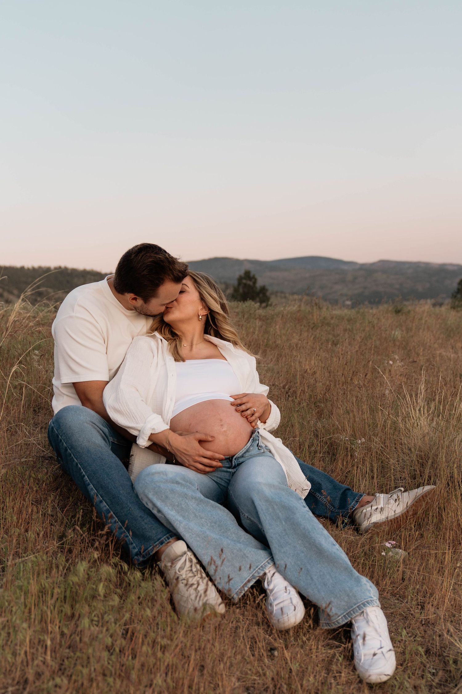 Couple sitting in a grassy field at sunset, man kissing pregnant woman's head while cradling her baby bump at Mount Munson in Penticton
