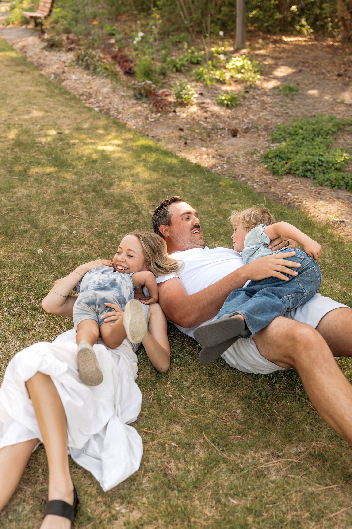 A joyful family laughing together on green grass in a sunny park, with two children cuddling their father.