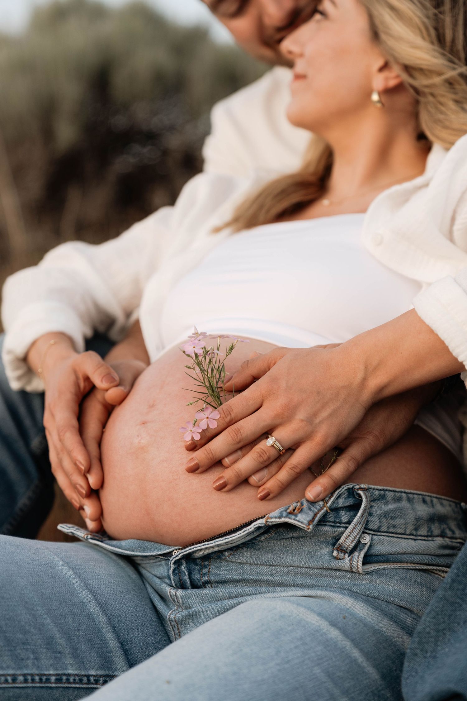 Close-up of a pregnant belly adorned with small pink flowers, with a couple embracing outdoors in Penticton