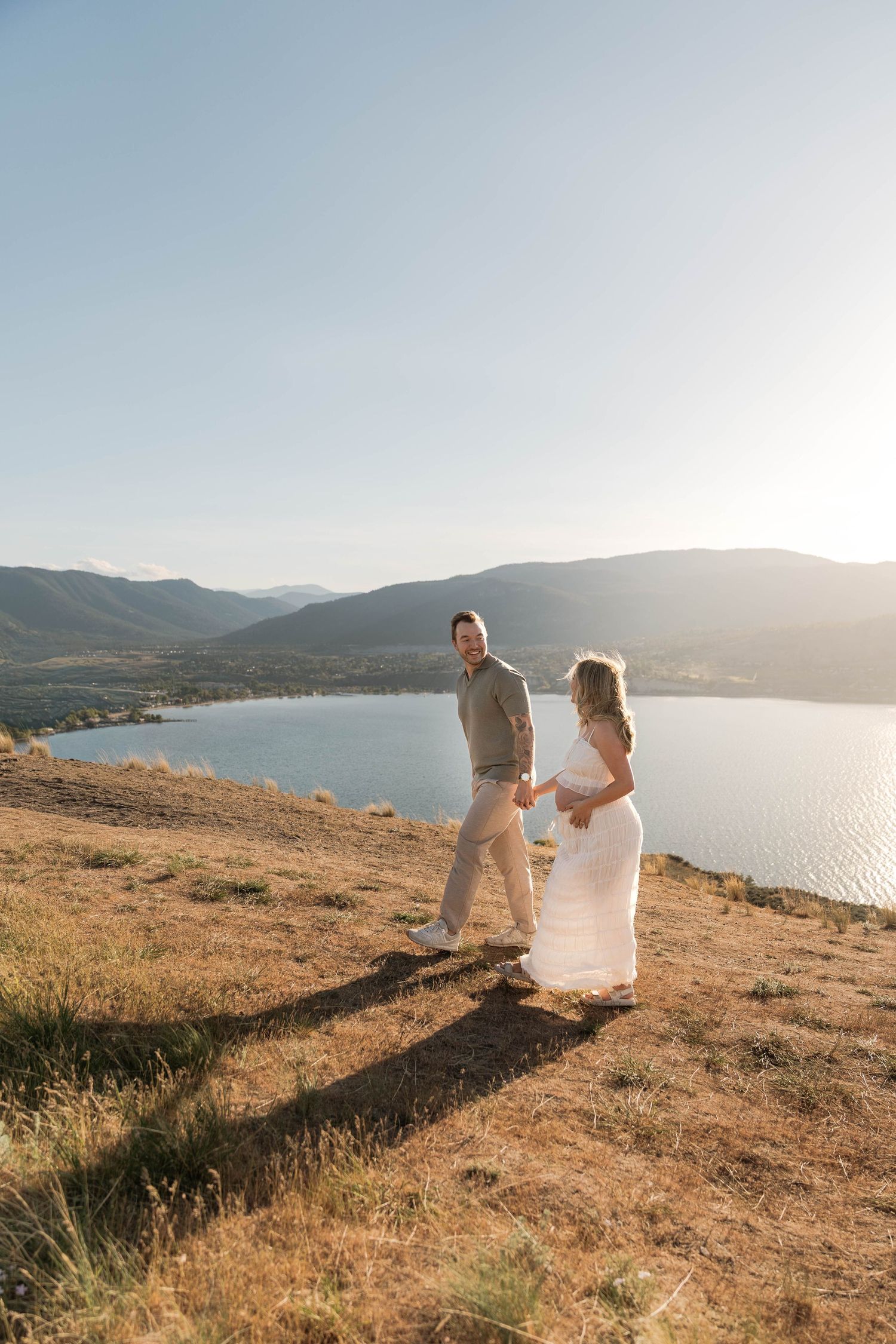 Couple standing together on a sun-drenched hillside with a stunning mountain lake backdrop at golden hour in Penticton