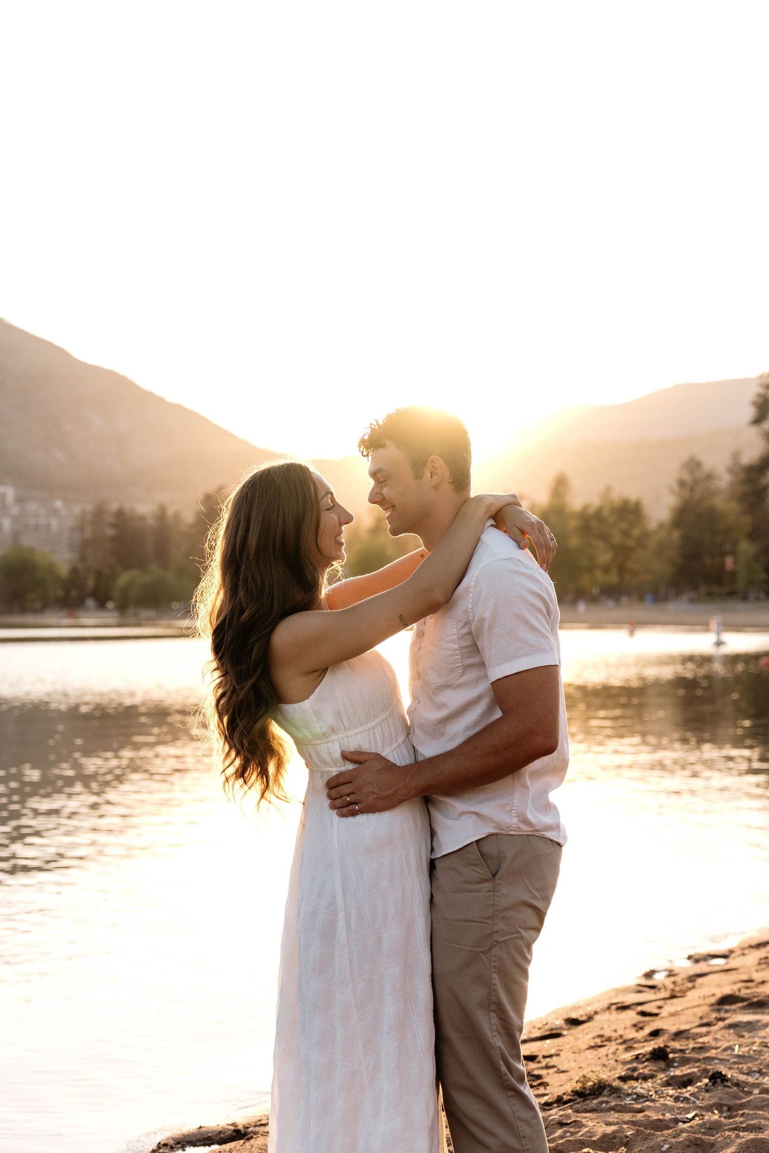 Couple embracing romantically at golden sunset by a lake with mountains in the background.