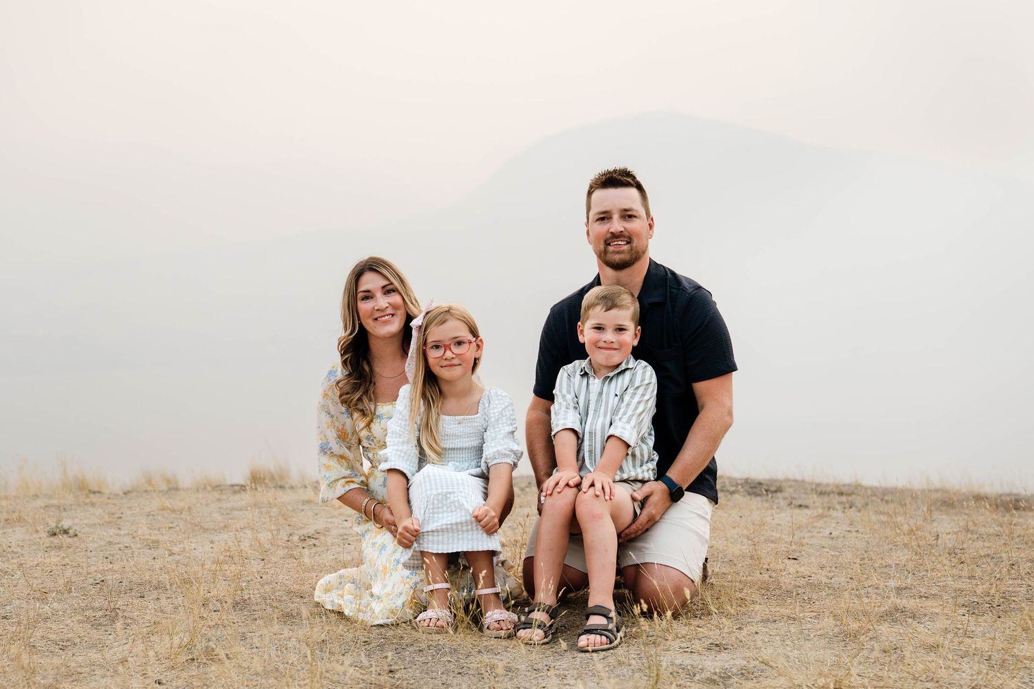 Family of four posing outdoors on dry grass with misty mountains in the background on a hazy day.