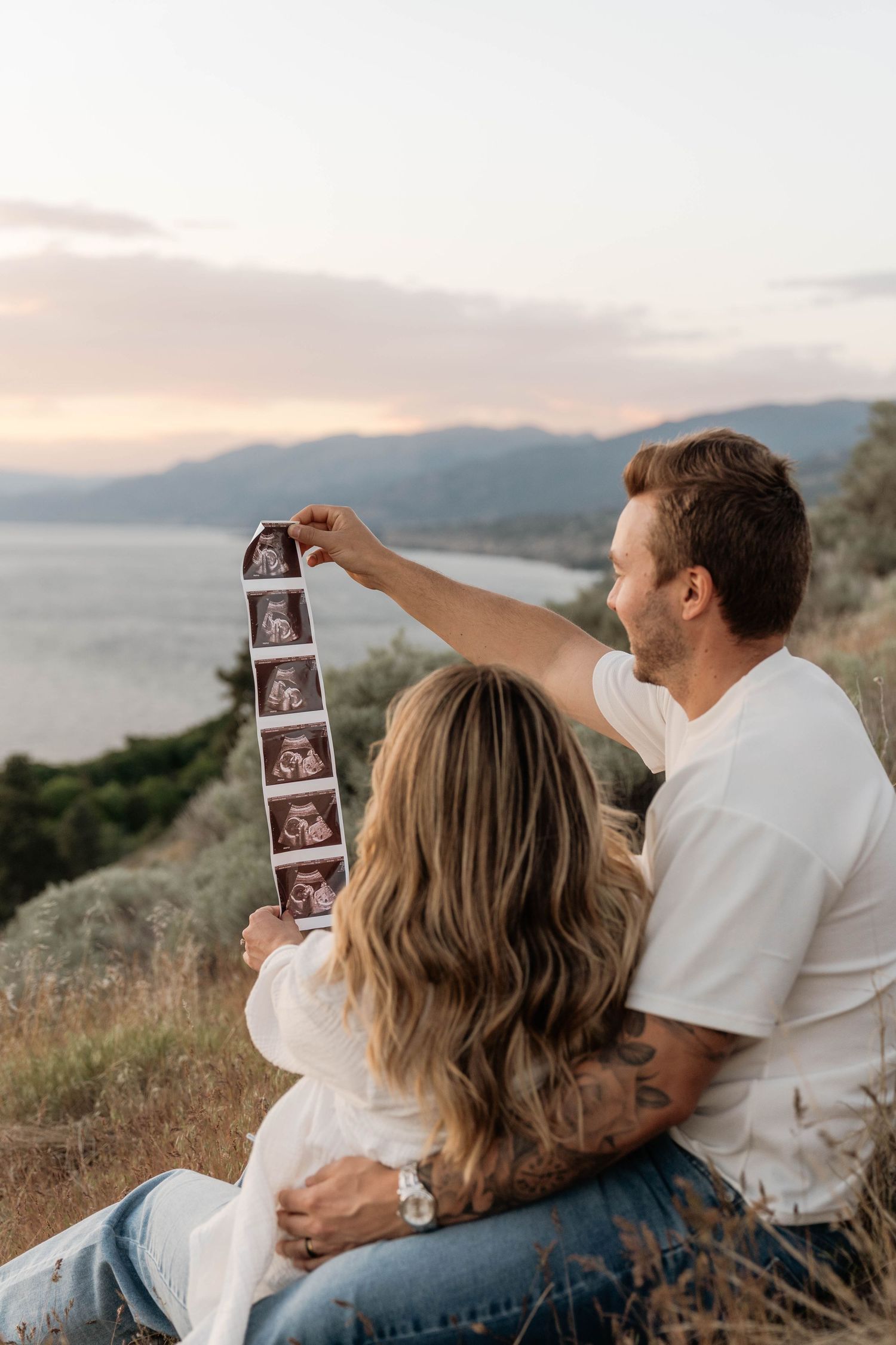 Couple sitting outdoors holding ultrasound photos with scenic mountain and lake view at sunset in Penticton