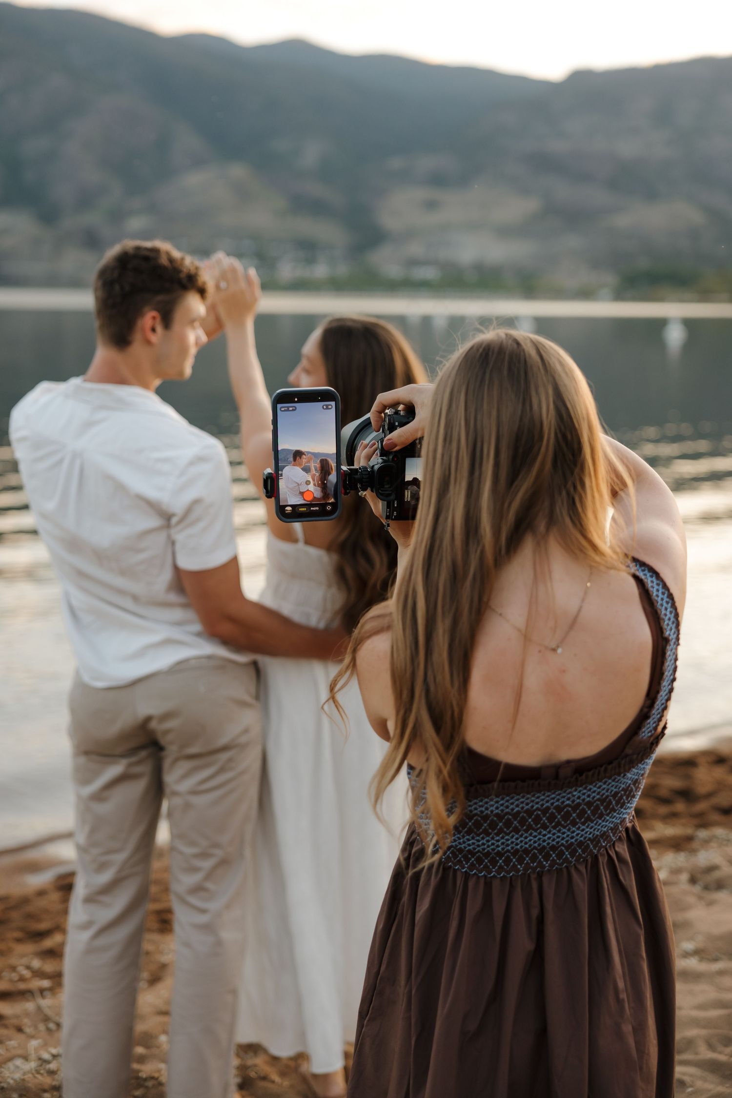 A photographer films a couple dancing near a lakeside at golden hour during an outdoor photo session in Penticton
