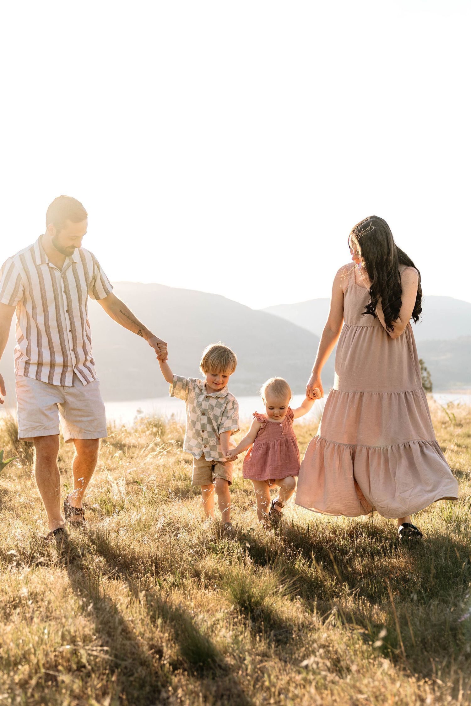 A family of four holds hands while walking through a golden meadow at sunset near a scenic lake in Penticton