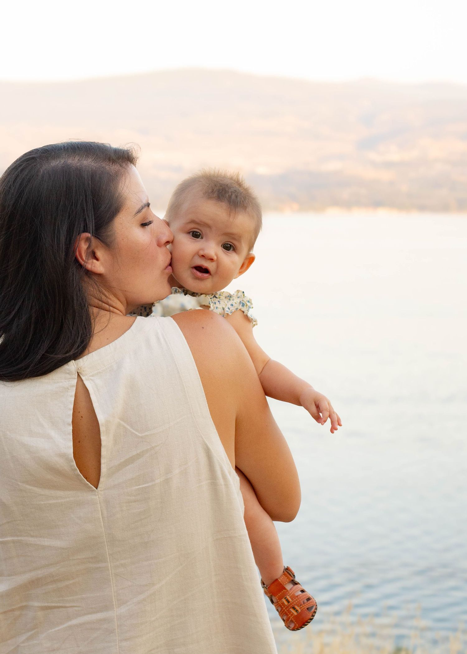 A dark-haired mother tenderly kisses her smiling baby on the cheek near a serene lake at sunset in Penticton