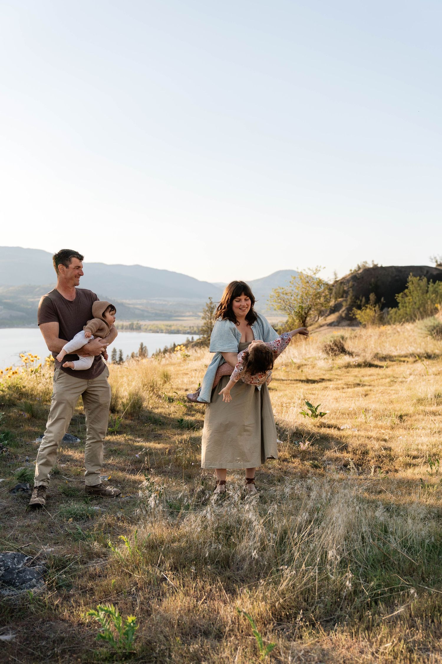 A family with two young children enjoys a sunny outdoor session on a hillside overlooking a peaceful lake in Penticton