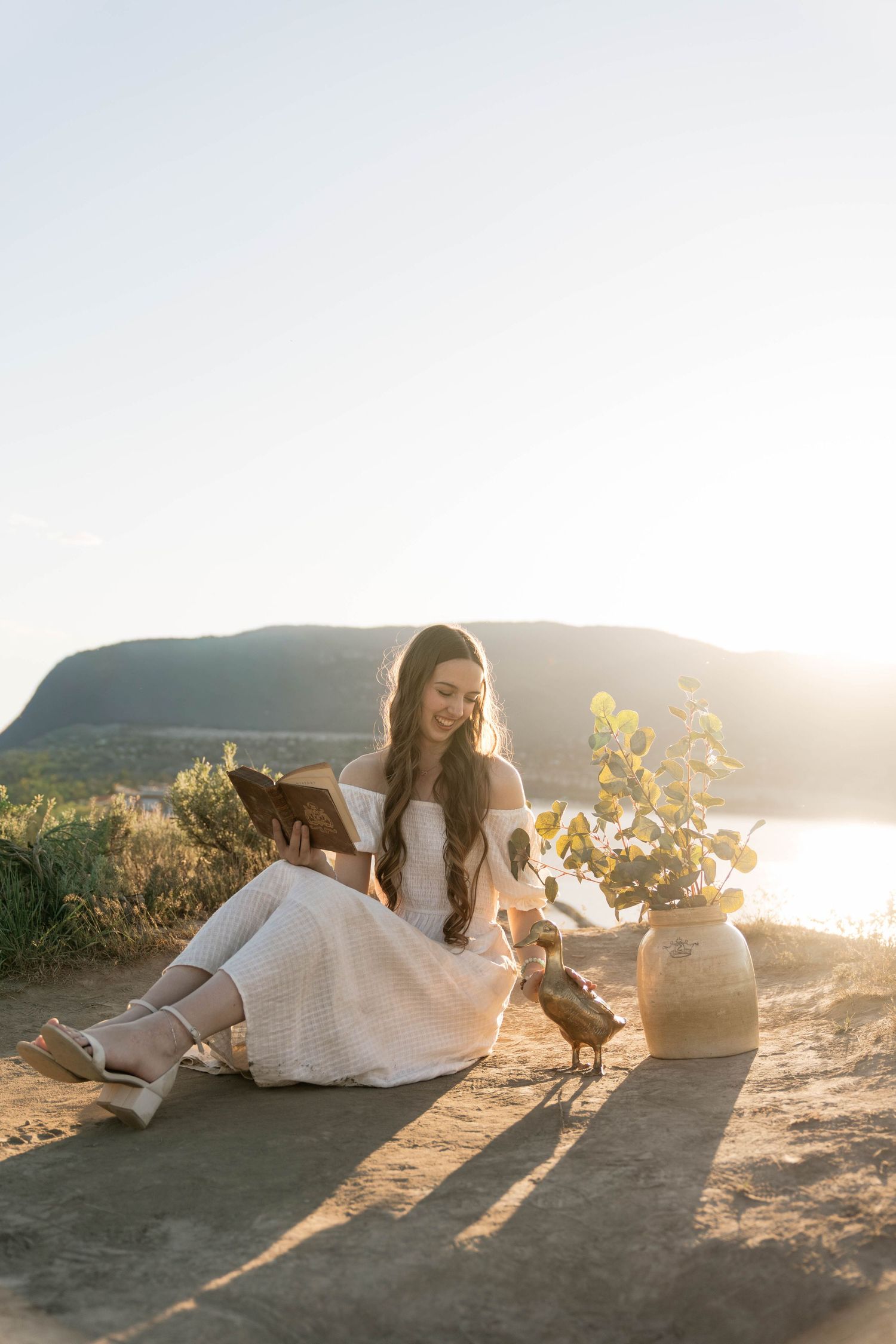 Woman in white dress reading a book outdoors at golden hour with a scenic lake and mountain backdrop.