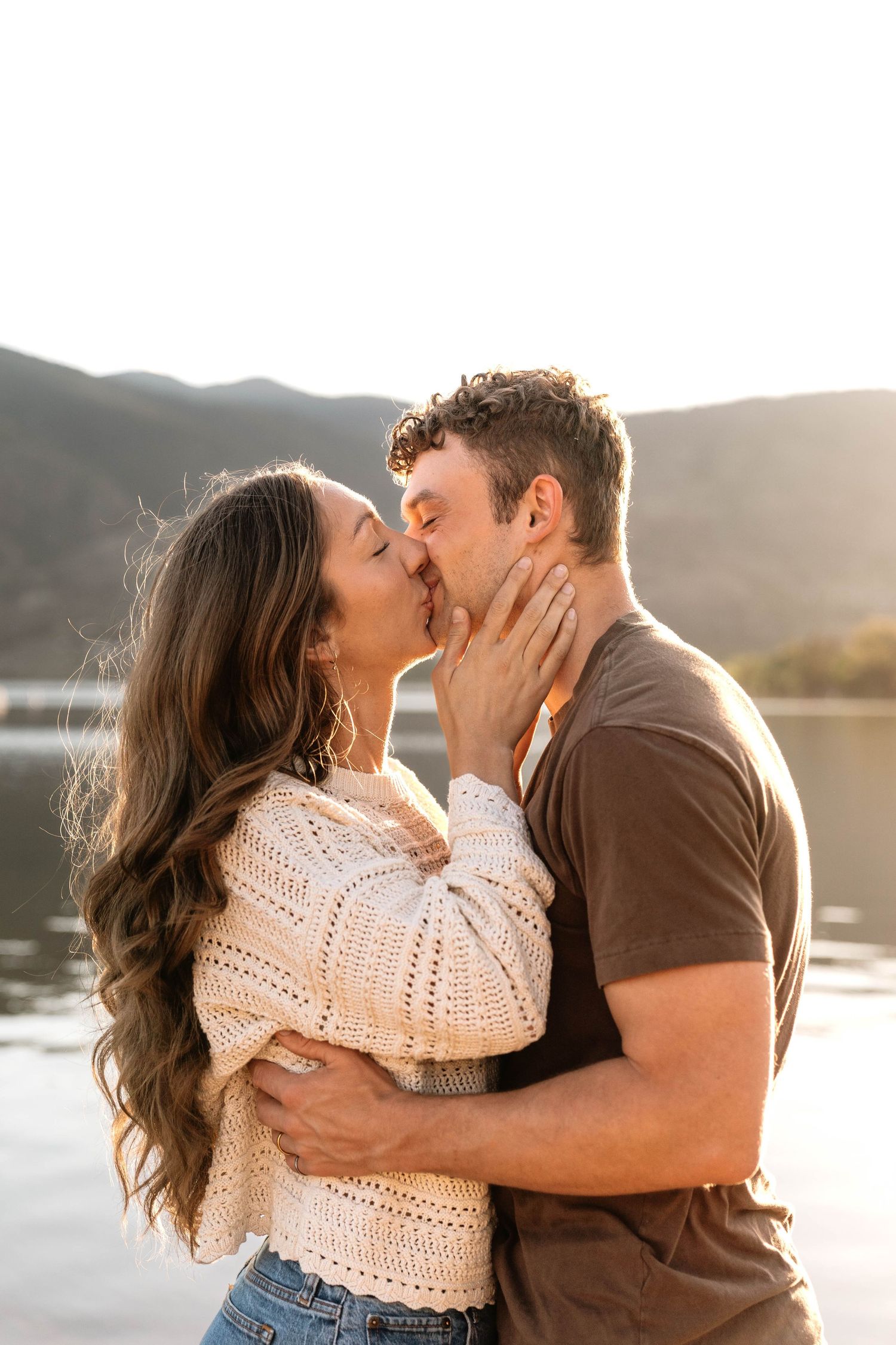 A couple shares a romantic kiss by a scenic mountain lake at golden hour, the woman in a cream sweater.