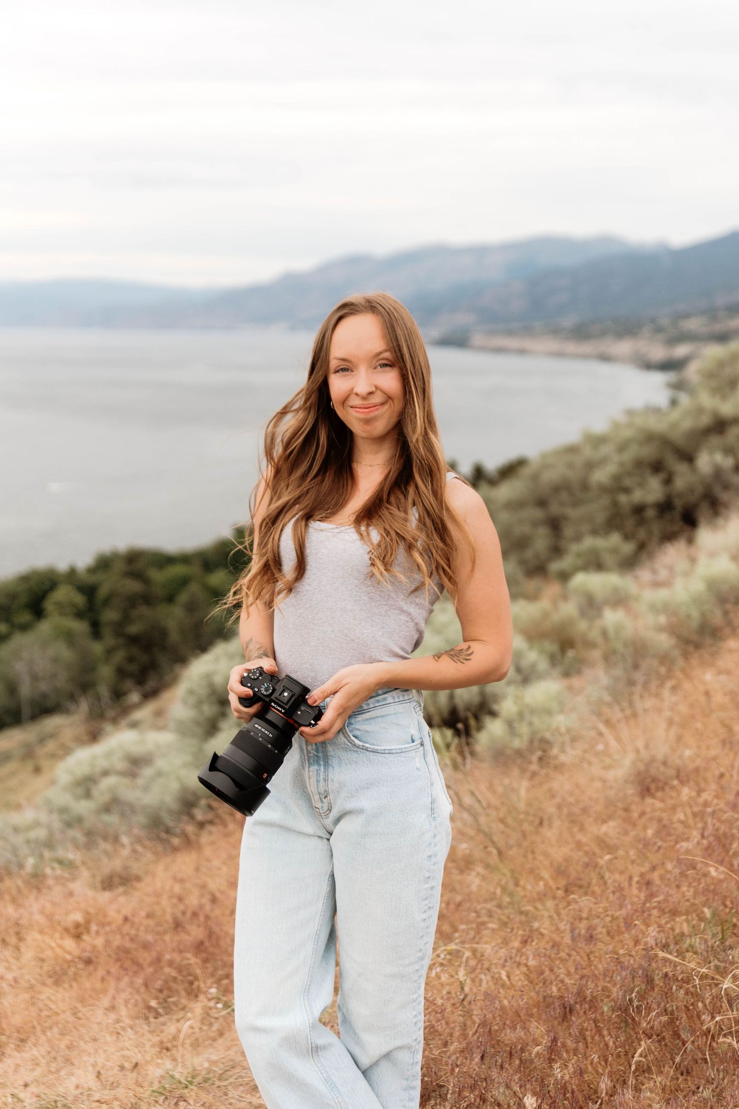 A smiling female photographer holds a camera while standing on a hillside with a lake and mountains behind her in Penticton