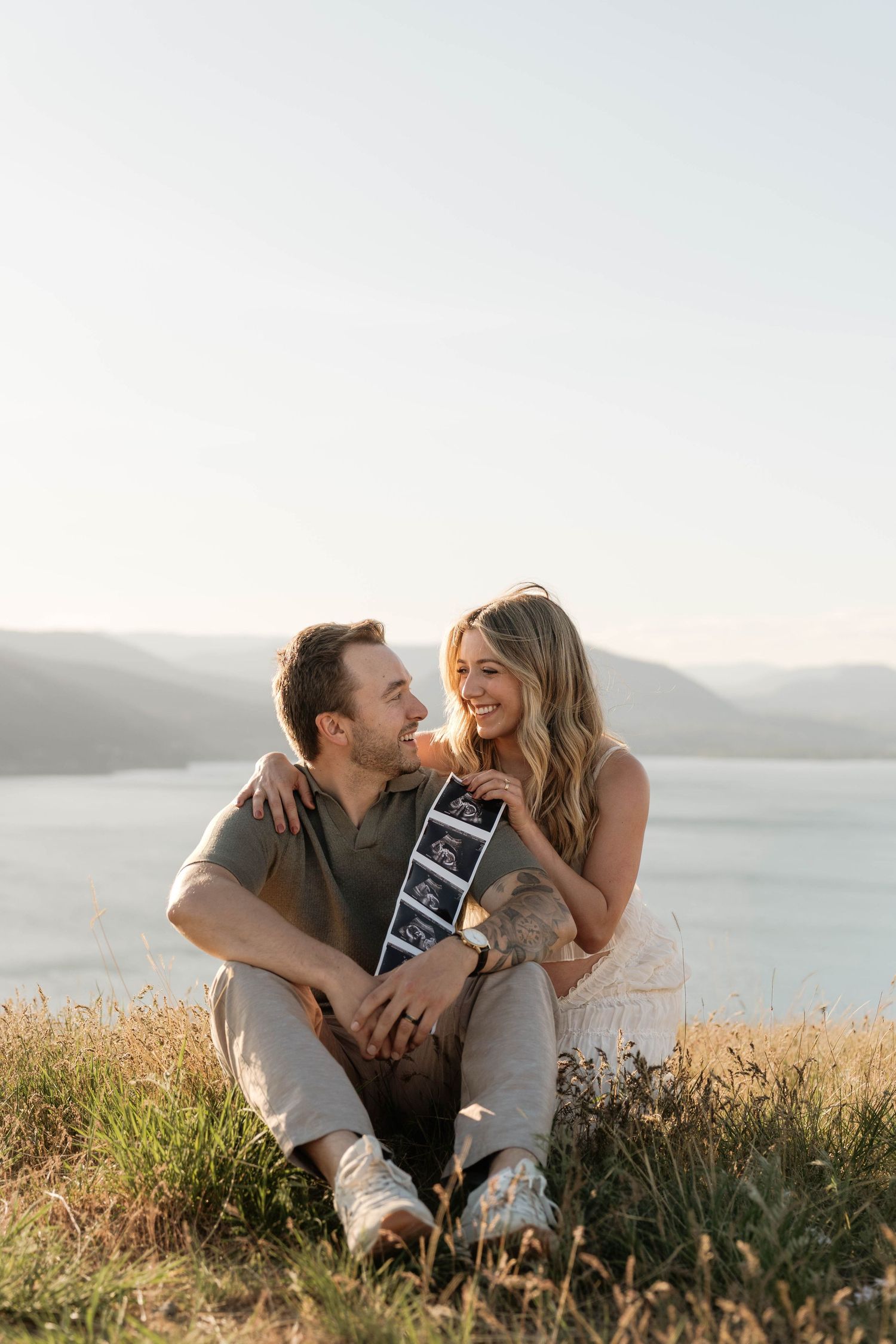 Smiling couple sitting on a hillside holding an ultrasound photo with a scenic lake and mountains in the background.