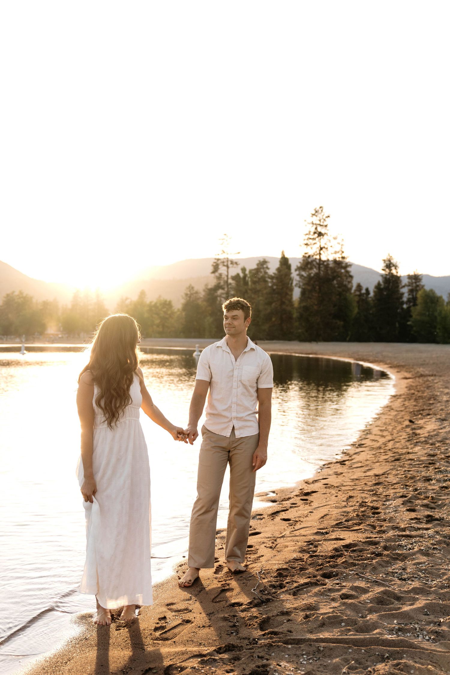 Couple holding hands on sandy lakeside beach at golden sunset with pine trees and mountains in background.