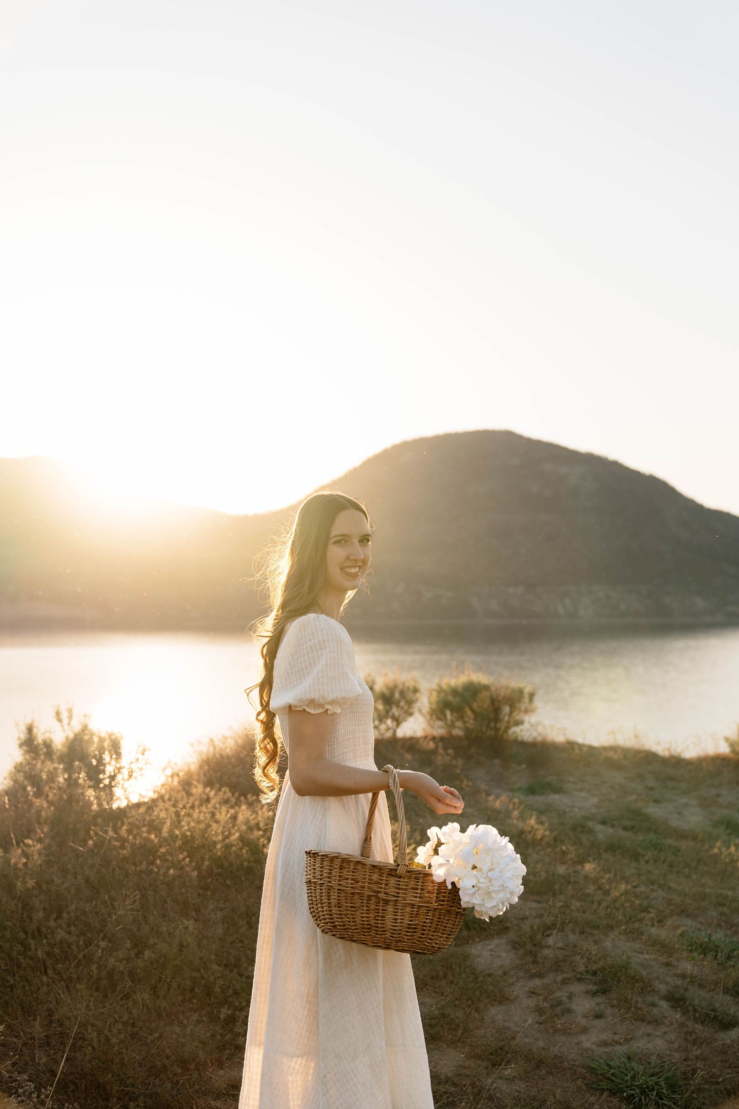 Smiling woman in white dress holding wicker basket with white flowers at golden hour near lake and hill.