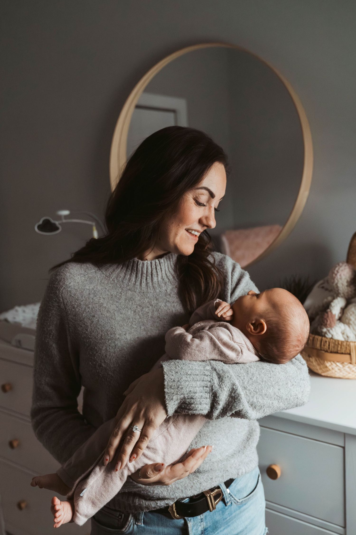 A smiling dark-haired woman in a gray sweater tenderly holds a newborn baby in a cozy nursery room.