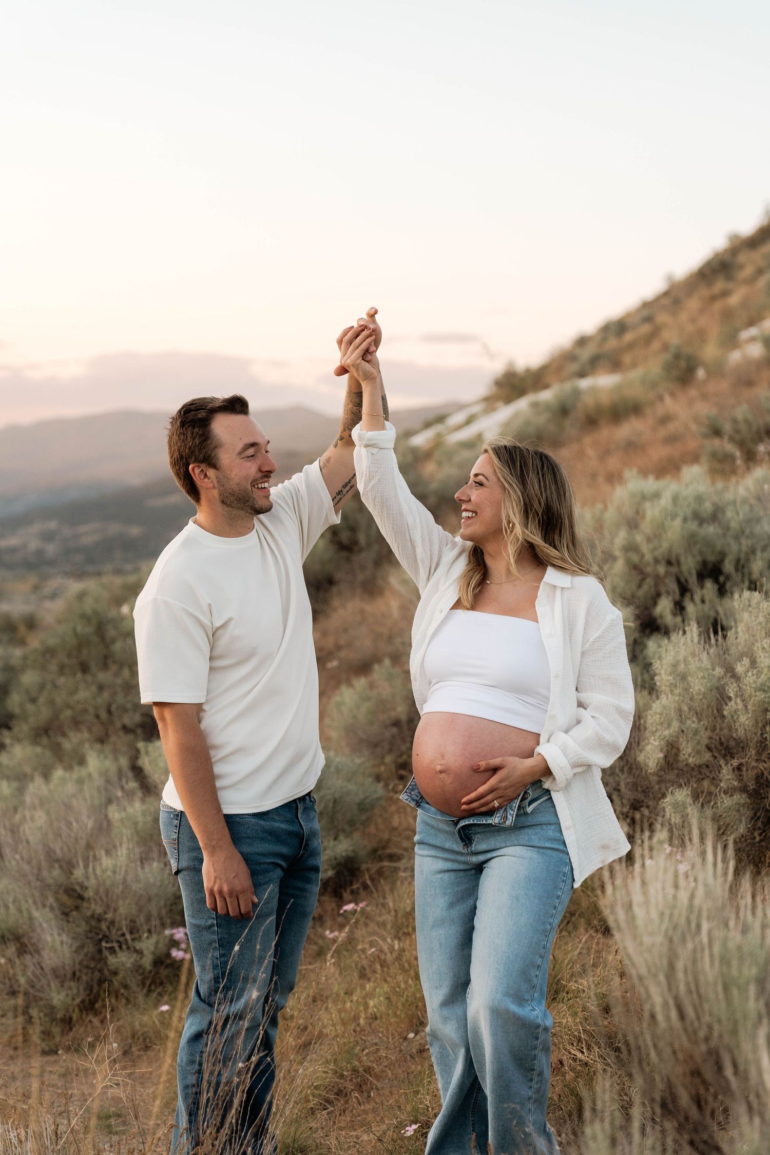 Happy pregnant couple holding hands raised outdoors at sunset, woman cradling baby bump, wearing white tops and jeans.