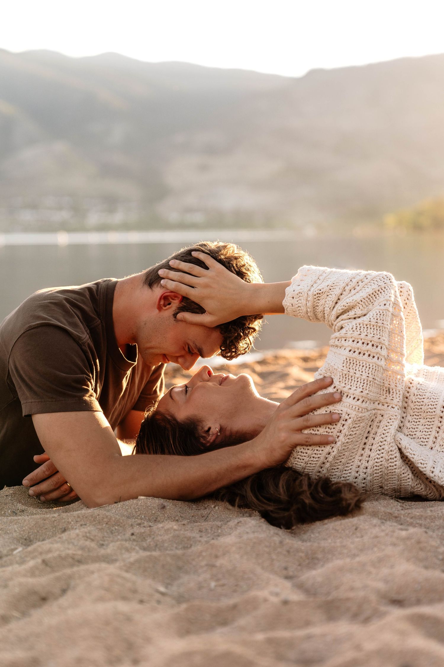 Couple sharing a romantic moment on a sandy beach at sunset with mountains and a lake in the background.