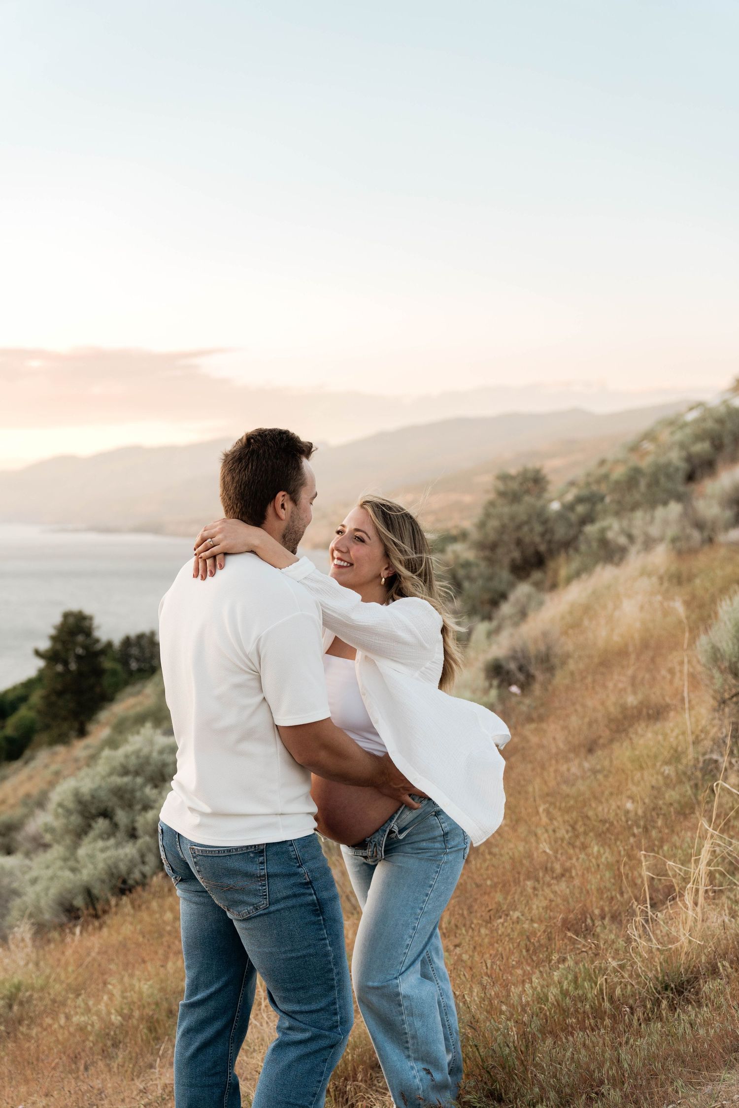 Laughing pregnant woman embraced by partner on a grassy hillside with a mountain lake at sunset in Penticton